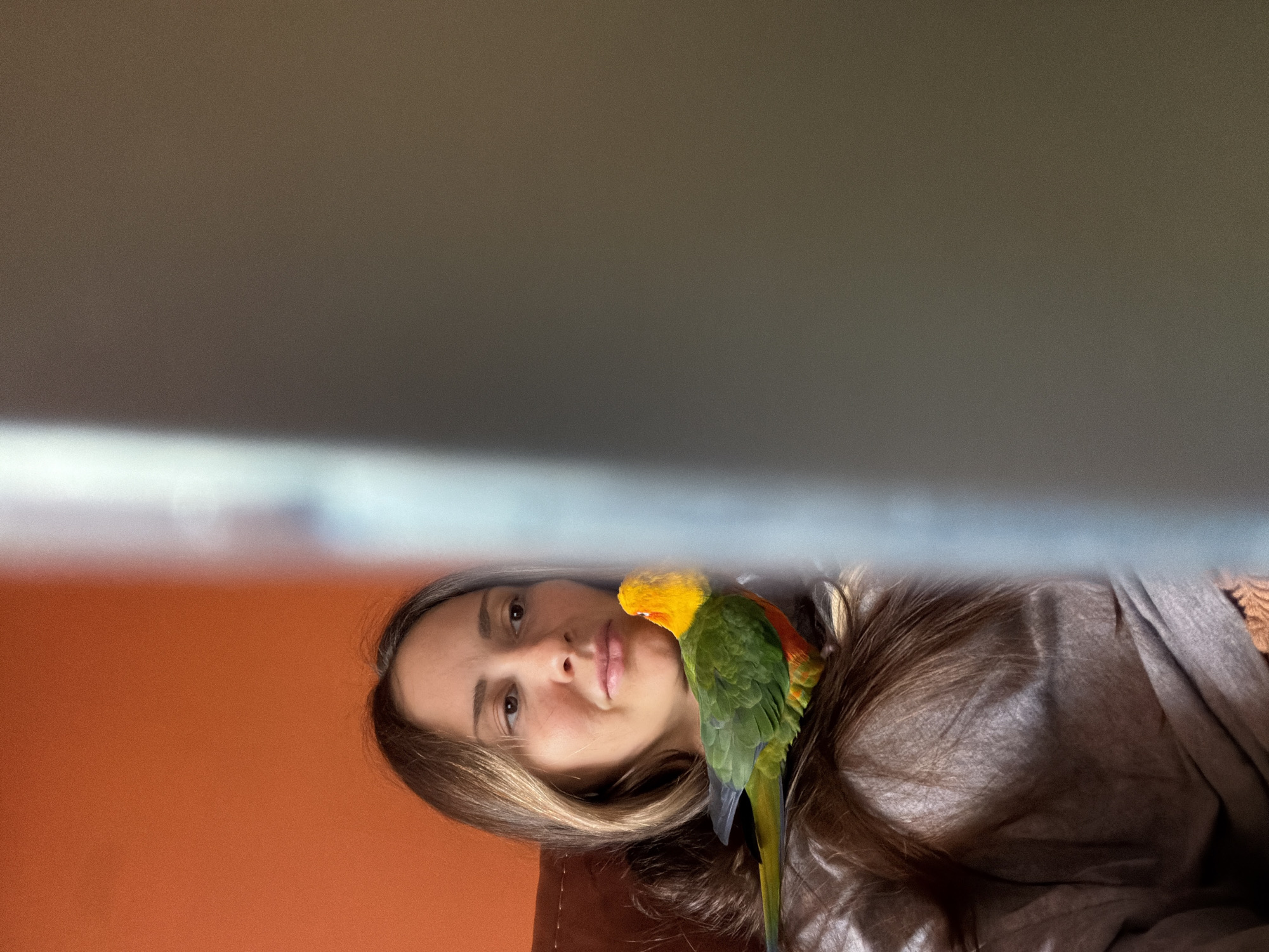 A woman with light brown hair rests her head on a brown pillow, with a Sun Conure perched on her shoulder. The bird has a yellow head, green body, and a long tail. The background consists of a blurred orange wall and a grey-toned ceiling or shelf. There is no evidence or indication of this image being located in the city of Khimki, Russia.