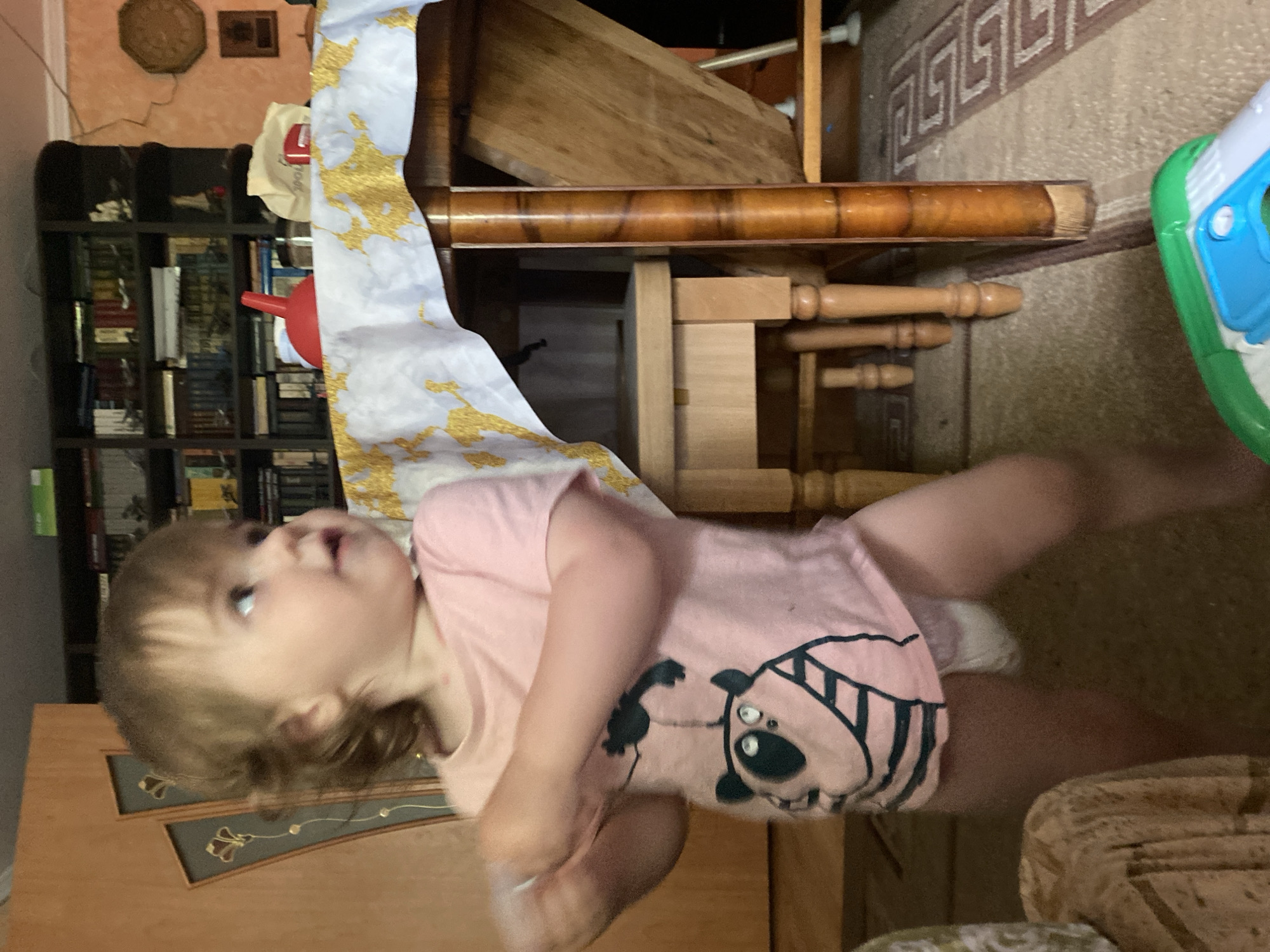 A young Caucasian child, appearing to be a toddler, is standing indoors in front of a bookshelf. The child is wearing a pink t-shirt with a cartoon panda graphic and a diaper. The child's hair is light brown and styled in a shaggy cut. The child is looking up and to the right of the frame. In the background, there is a wooden bookshelf filled with books and decorative items. A table draped with a white and gold patterned cloth is also visible, as is a portion of a rug with a Greek key pattern. A colorful toy is on the floor in the foreground. The lighting suggests an indoor setting during the daytime.