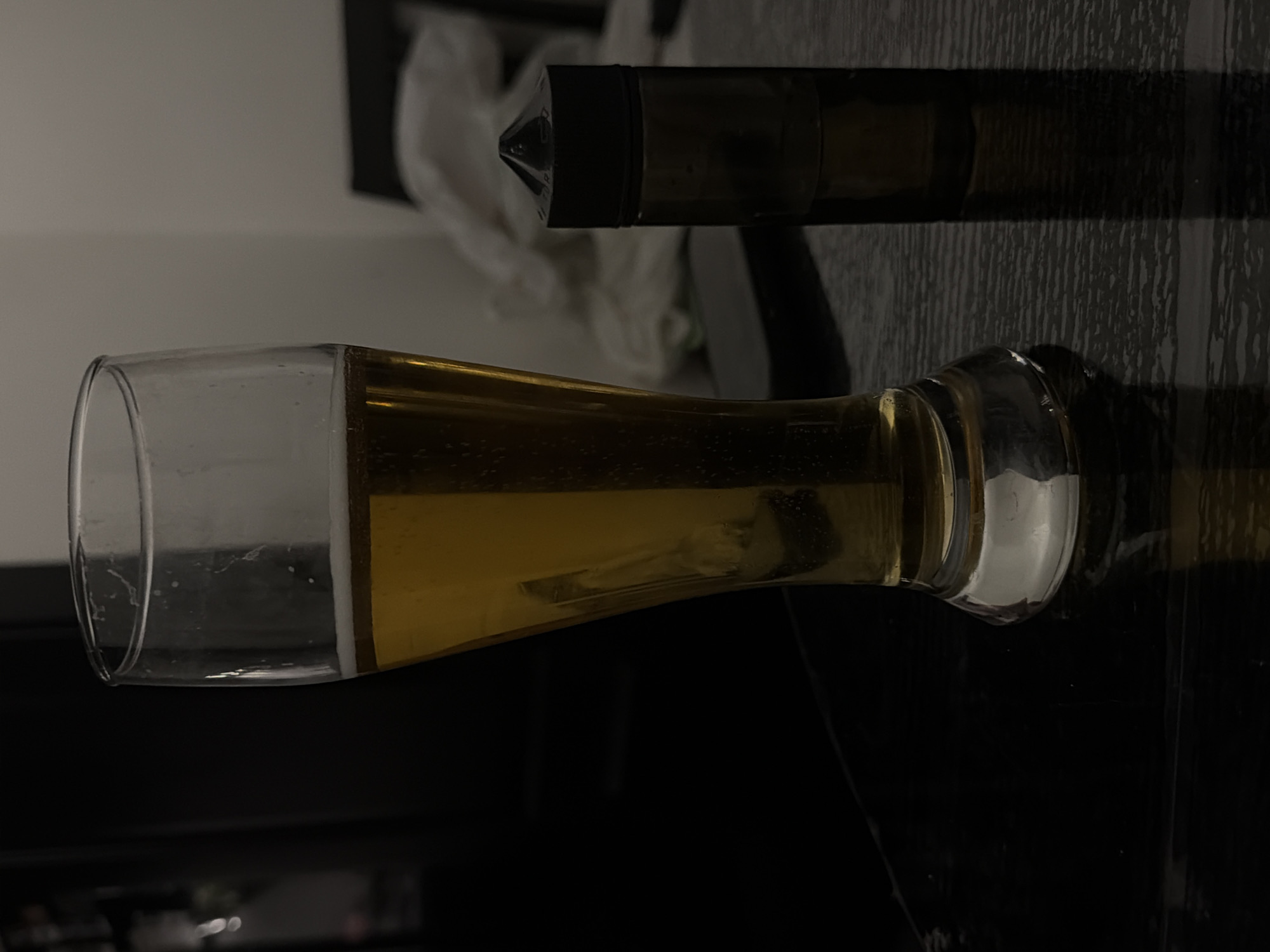 A glass of beer and a small bottle with a black cap are placed on a dark, reflective surface. The glass is tall and has a slight curve, filled with golden-colored liquid and a frothy head. The bottle is dark brown and opaque, with a pointed black cap. The background is dimly lit, with a light-colored wall on the right and a dark, out-of-focus area on the left. The scene appears to be indoors. There is no indication of the city of Bila Tserkva, Ukraine.