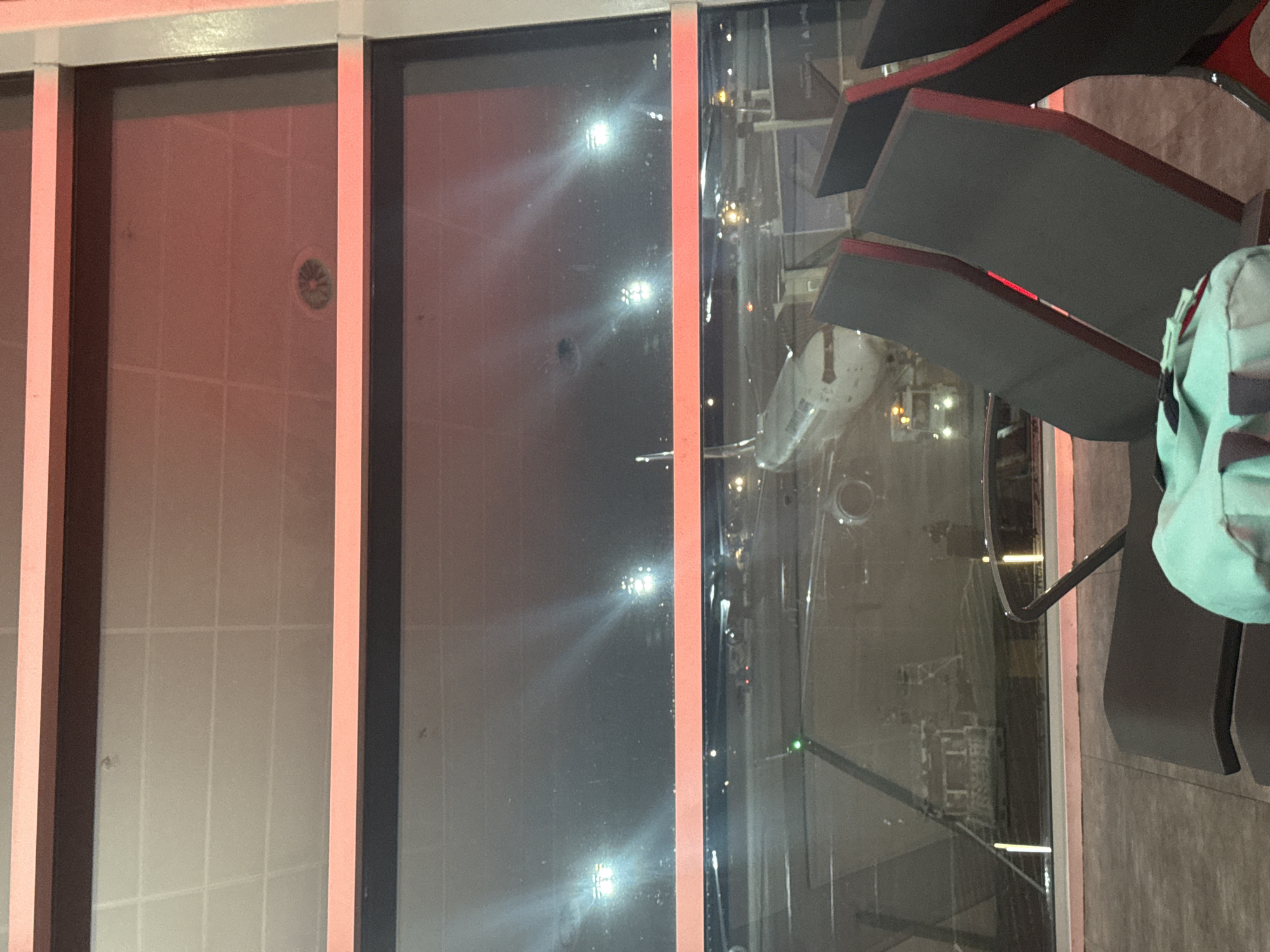The image depicts an airplane on a tarmac viewed through a glass window, likely in an airport terminal. The interior includes seating with gray and red details, and a light blue backpack is visible on one of the seats. The window frames are dark gray with light red accents. The tarmac scene is blurred due to the reflections on the glass. The location is likely Tbilisi International Airport in Tbilisi, Georgia, given the context of the request.