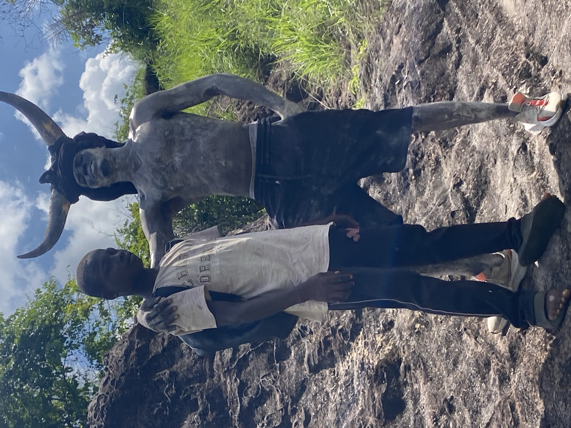 A man with dark skin stands in front of a statue of a man with horns, in Afikpo, Nigeria. The statue is of a muscular man with two large horns protruding from his head. The statue has on shorts and shoes. The man in front of the statue is wearing a white T-shirt, dark pants, and sandals. He has his arms around the statue. In the background, there is lush vegetation and a sky with clouds. The ground and area around them appears to be rocky terrain.