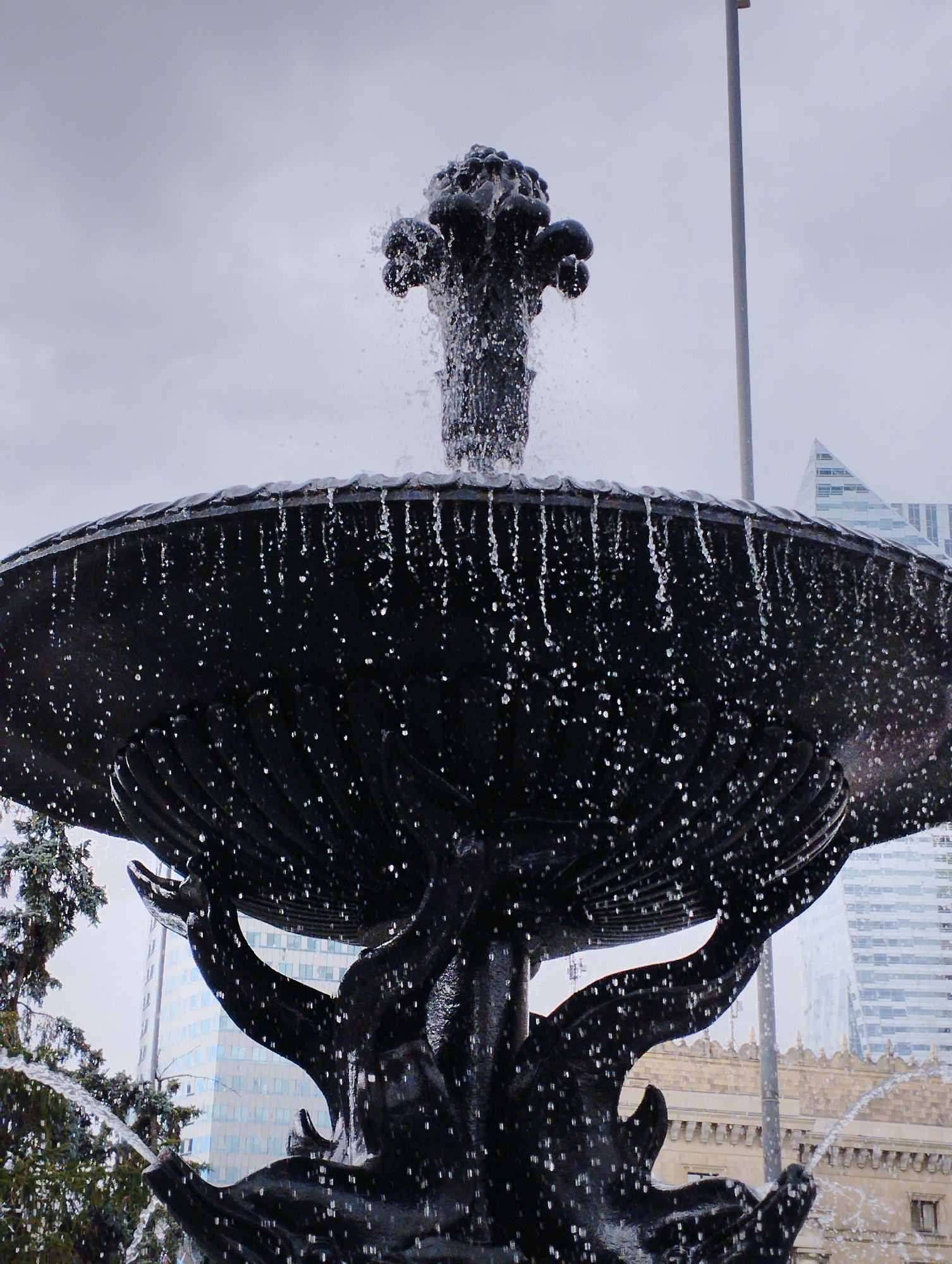 The image shows a close-up, low-angle view of a dark, ornate fountain with water spraying from its top and cascading down its tiers. The fountain appears to be made of cast iron or a similar dark metal, with intricate sculpted details. Water droplets are visible in the air and on the fountain's surfaces. In the background, blurred modern buildings, including a tall skyscraper with a distinctive triangular top, are visible under a cloudy sky. The fountain is likely located in a public square or park in Warsaw, Poland, given the presence of contemporary architecture common in the city. The scene is dynamic due to the flowing water, and the overall mood is somewhat muted due to the overcast weather. There are no people visible in the frame.