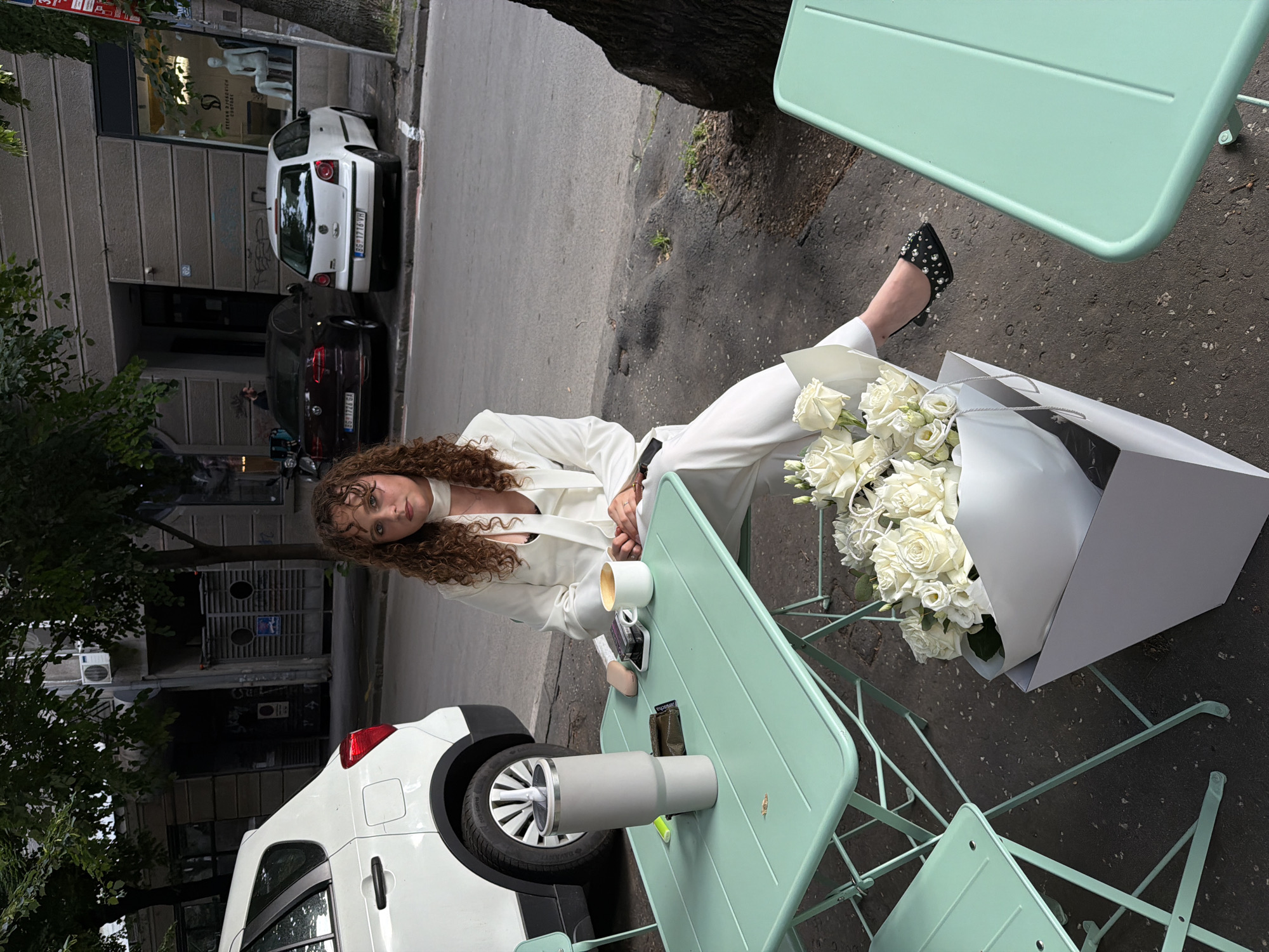 A young, curly-haired woman wearing a white pantsuit sits at a light-green metal bistro table on a street in Belgrade, Serbia. A large bouquet of white roses in a white paper cone sits beside her. On the table are a white coffee cup, a mobile phone, a small bag, and a tall, gray, insulated travel mug. The road in the background is busy. Cars are parked adjacent to the sidewalk. A white van is angled into the table and chairs. The background contains storefronts, trees, and utility poles.