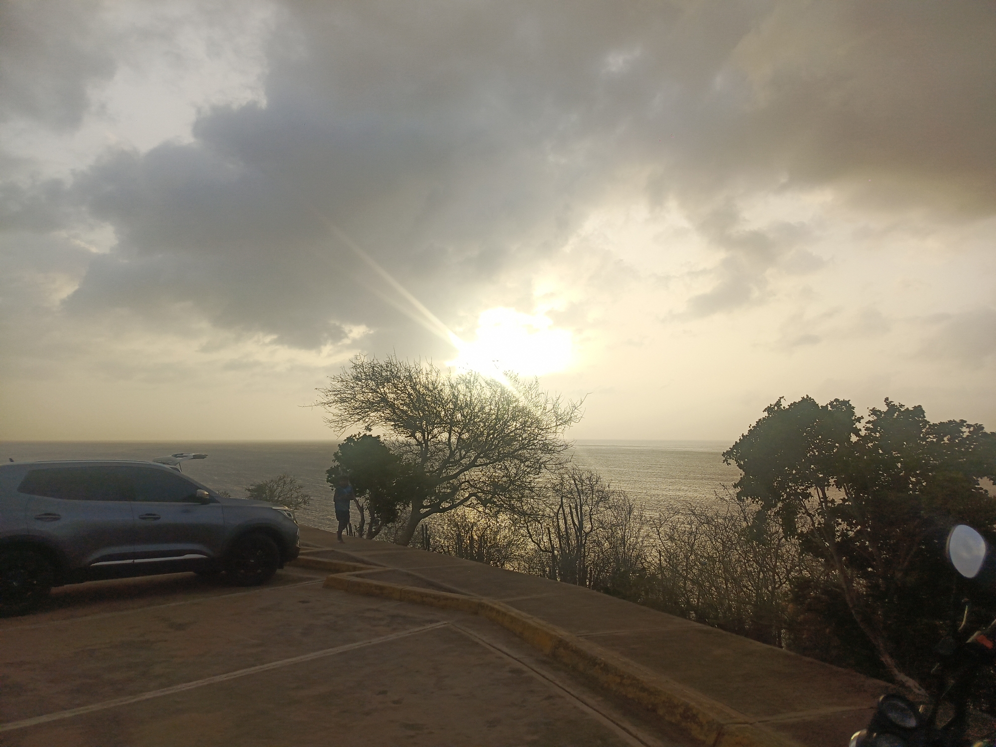 The image captures a dramatic coastal landscape at what appears to be sunset, overlooking the sea in Juan Griego, Venezuela. The scene is dominated by a cloudy sky where the sun breaks through with a bright, radiant burst, casting a warm, golden glow across the water and illuminating the clouds from behind.
In the foreground, a dark grey SUV is parked on an asphalt or concrete surface with yellow painted curbs. The vehicle is seen from its left side, facing slightly towards the right, with dark wheels prominent. To the right of the SUV, parts of a motorcycle, including a mirror and handlebar, are visible in the bottom right corner, suggesting the photo was taken from or near a motorbike.
The midground features several trees and shrubs, many of them bare or silhouetted against the bright sky and glistening sea, emphasizing their intricate branch patterns. One person is visible standing near the edge of the elevated viewpoint, seemingly looking out towards the ocean, their figure silhouetted by the strong backlighting. The individual appears to be wearing a blue shirt.
Beyond the trees, the vast expanse of the ocean stretches towards the horizon, reflecting the golden light of the sun. The water appears calm, with subtle ripples visible on its surface. The overall atmosphere is serene and vibrant, characteristic of a coastal sunset. No discernible text is visible in the image.