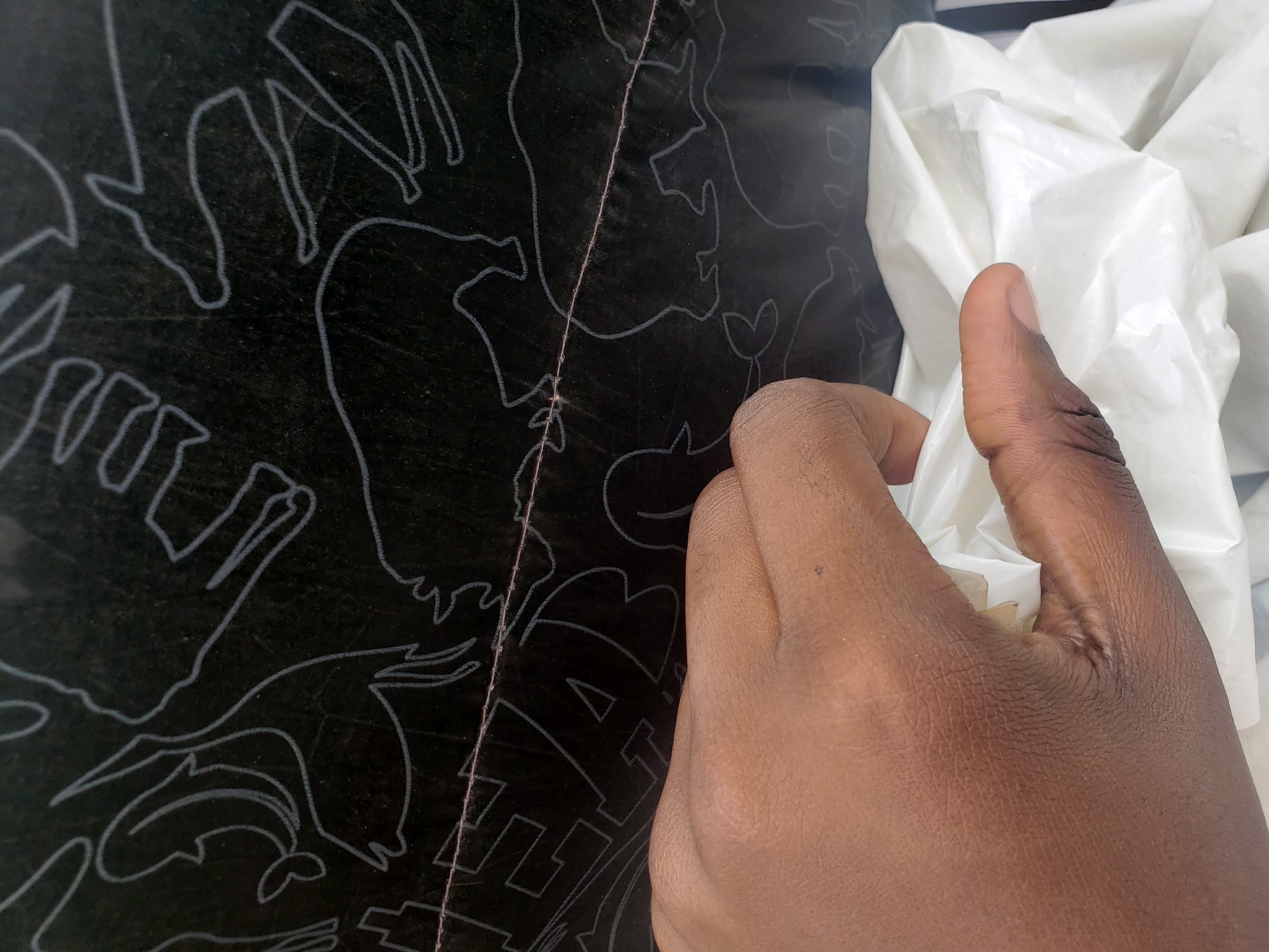 In Narayi, Nigeria, a close-up shot captures a person's hand, with dark skin and visible wrinkles, holding a crumpled piece of white material, possibly plastic. The hand is positioned in the lower half of the frame, with the fingers and thumb gripping the material. Behind the hand, a dark surface with a repeating pattern of white line drawings of animals is visible. A thin, horizontal pink line runs across this patterned surface, dividing it into upper and lower sections. The overall environment appears to be indoors, given the lighting and the texture of the dark surface.