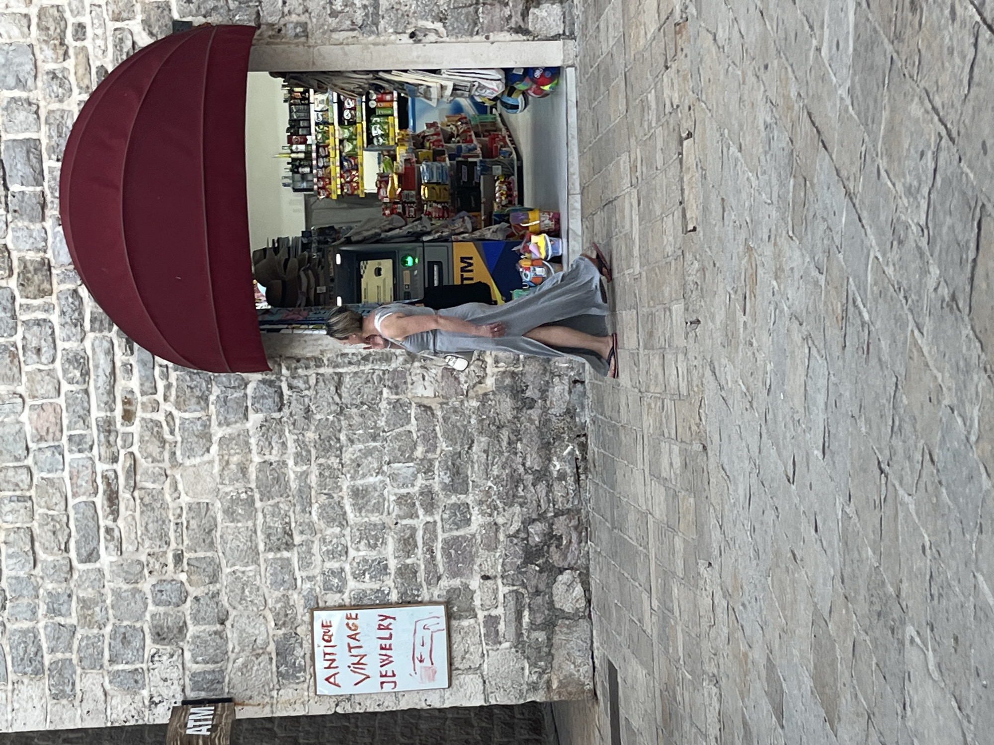 A woman in a long, grey, sleeveless dress and flip-flops walks past a stone building on a cobblestone street in Budva, Montenegro. She is carrying a white purse. To her left, a sign on the stone wall reads "ATM" in black text on a wooden background. Below that, a white sign with red lettering says "ANTIQUE VINTAGE JEWELRY" and has a small red drawing of an arm. To the woman's right, an open storefront with a red awning displays shelves stocked with various goods, including bottles and packaged items. An ATM machine is visible inside the store. The sky is not visible.