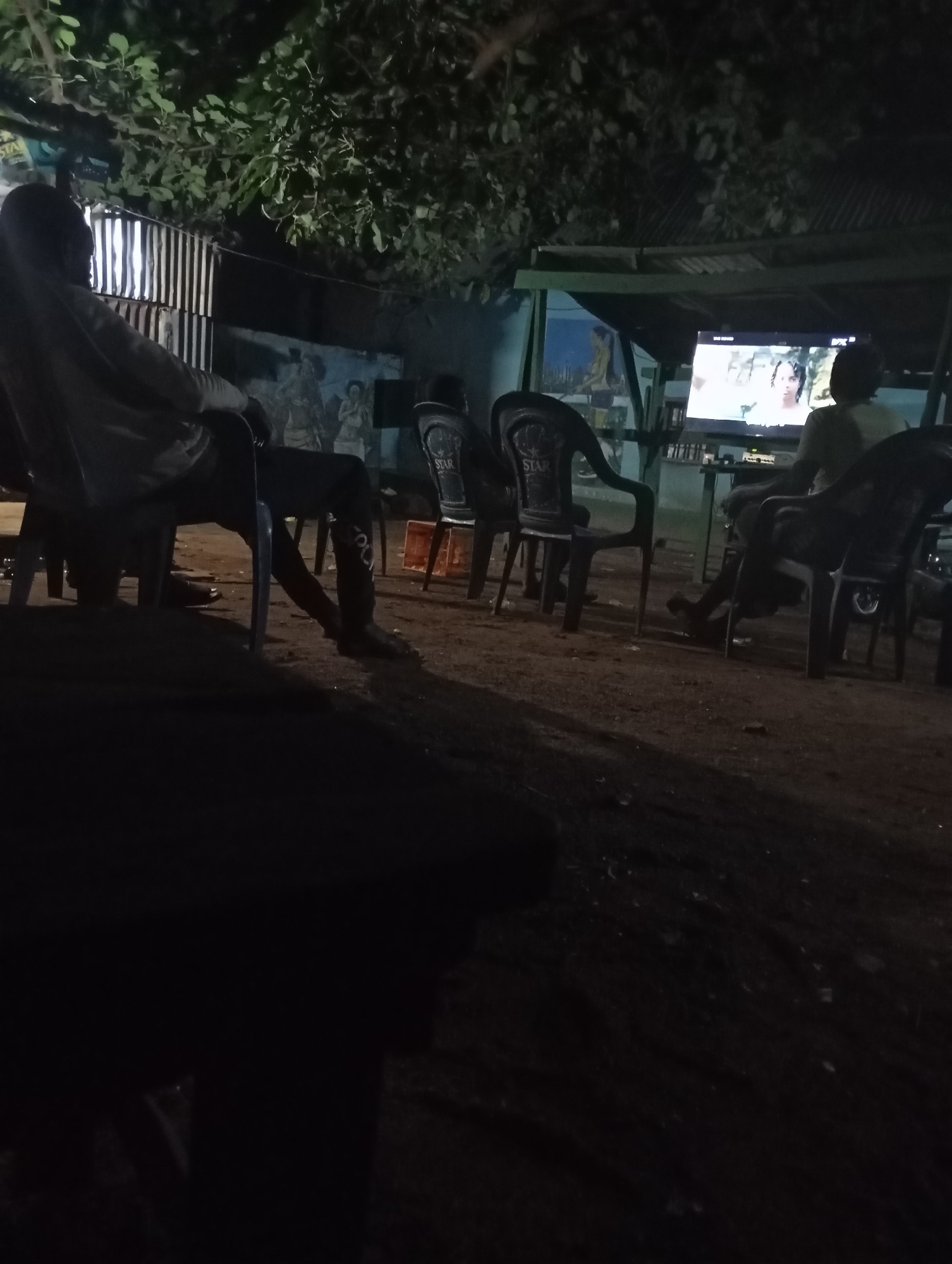 In the city of Unknown, Unknown, at night, a group of people are gathered outdoors to watch television. The scene appears to be in a somewhat informal setting, possibly a street or a community area, with a corrugated metal fence in the background. Several individuals are seated on plastic chairs, facing a television screen that is displaying a program. The lighting is dim, with the television screen providing a primary light source. Some of the chairs are visible with "STAR" branding. The ground is dirt.