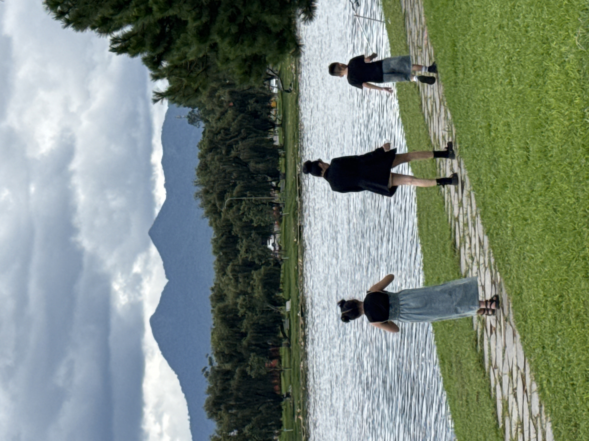 The image captures a scenic view of Xuan Huong Lake in Da Lat, Vietnam, under a cloudy sky. Three individuals, two children and one adult, are walking on a stone pathway adjacent to the lake. The adult, dressed in a black coat and shorts, walks ahead of a young boy wearing a black t-shirt and shorts. A young girl in a blue dress walks on the pathway further ahead, facing away from the camera. The lake's surface is rippled, reflecting the overcast sky and the lush green trees lining the shore. In the background, mountains rise, shrouded in mist. The scene depicts a leisurely stroll by the lake.