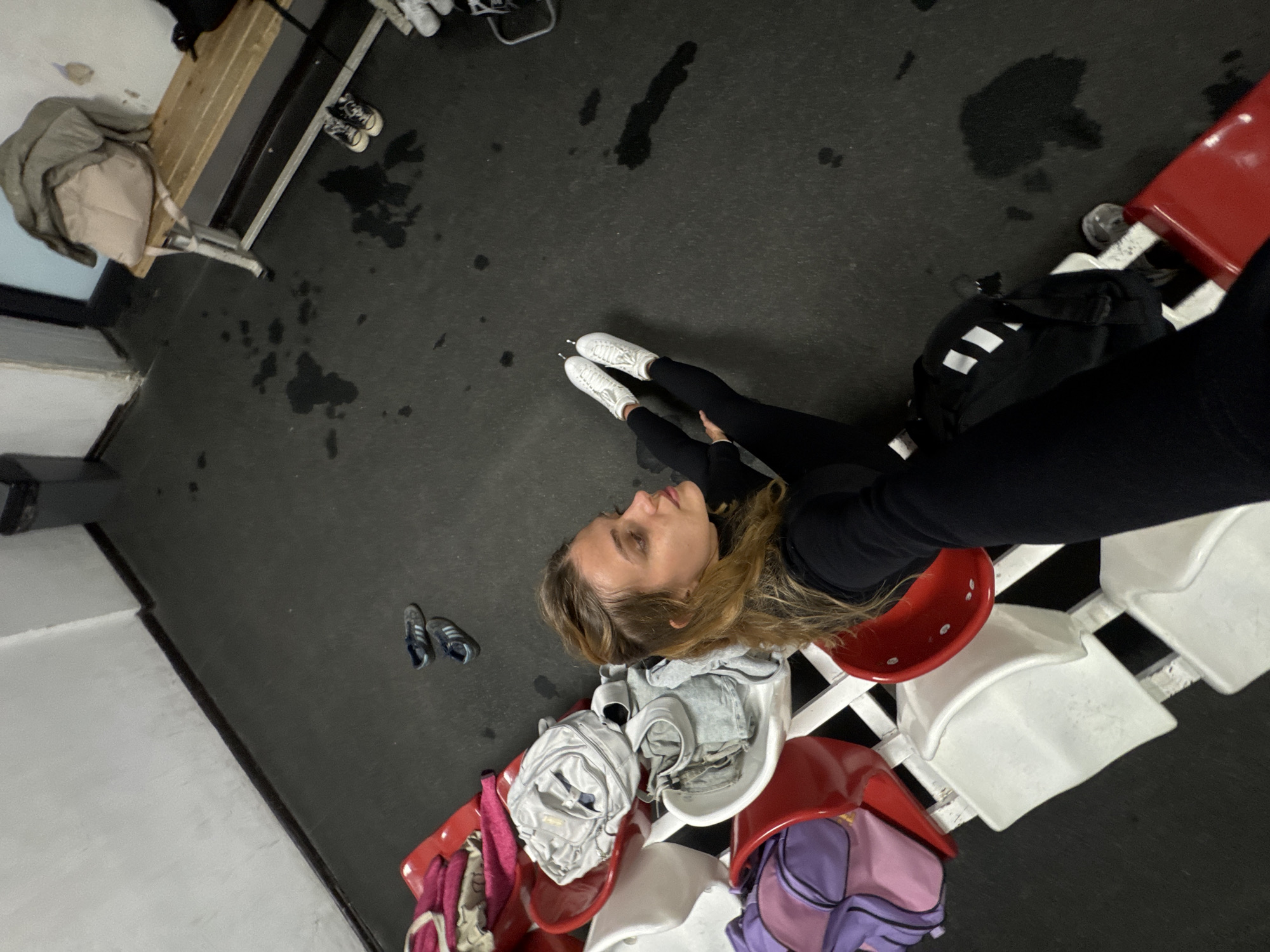 A woman is sitting in a stadium in Unknown, Unknown. She is wearing white skates and black leggings, and she has blonde hair. She is looking to the left and appears to be taking a selfie. There are red and white stadium seats behind her, and a black backpack with white stripes is sitting on the seat to her right. There are water puddles on the ground, and a bench is visible on the left. There is a gray jacket and a pair of black and white sneakers on the bench.