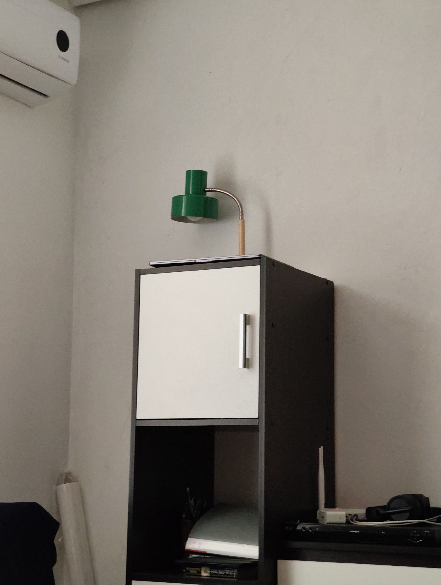 The image shows an interior scene, likely a room, with a white wall and a grey floor. In the center of the frame, a dark brown and white cabinet is visible. The top part of the cabinet has a white door with a silver handle. On top of this cabinet, a green desk lamp with a silver arm is positioned, casting a shadow on the wall. To the left of the cabinet, a white air conditioning unit is mounted on the wall. The bottom part of the cabinet is open and contains some items, including what appears to be a binder and a black object. On the shelf below, there is a white router with an antenna, headphones, and some electronic devices. To the left of the cabinet, a rolled-up white object, possibly a poster or blueprint, is leaning against the wall. The overall lighting suggests it is daytime. There are no people visible in the image. The image is located in Komsomolsk-on-Amur, Russia.