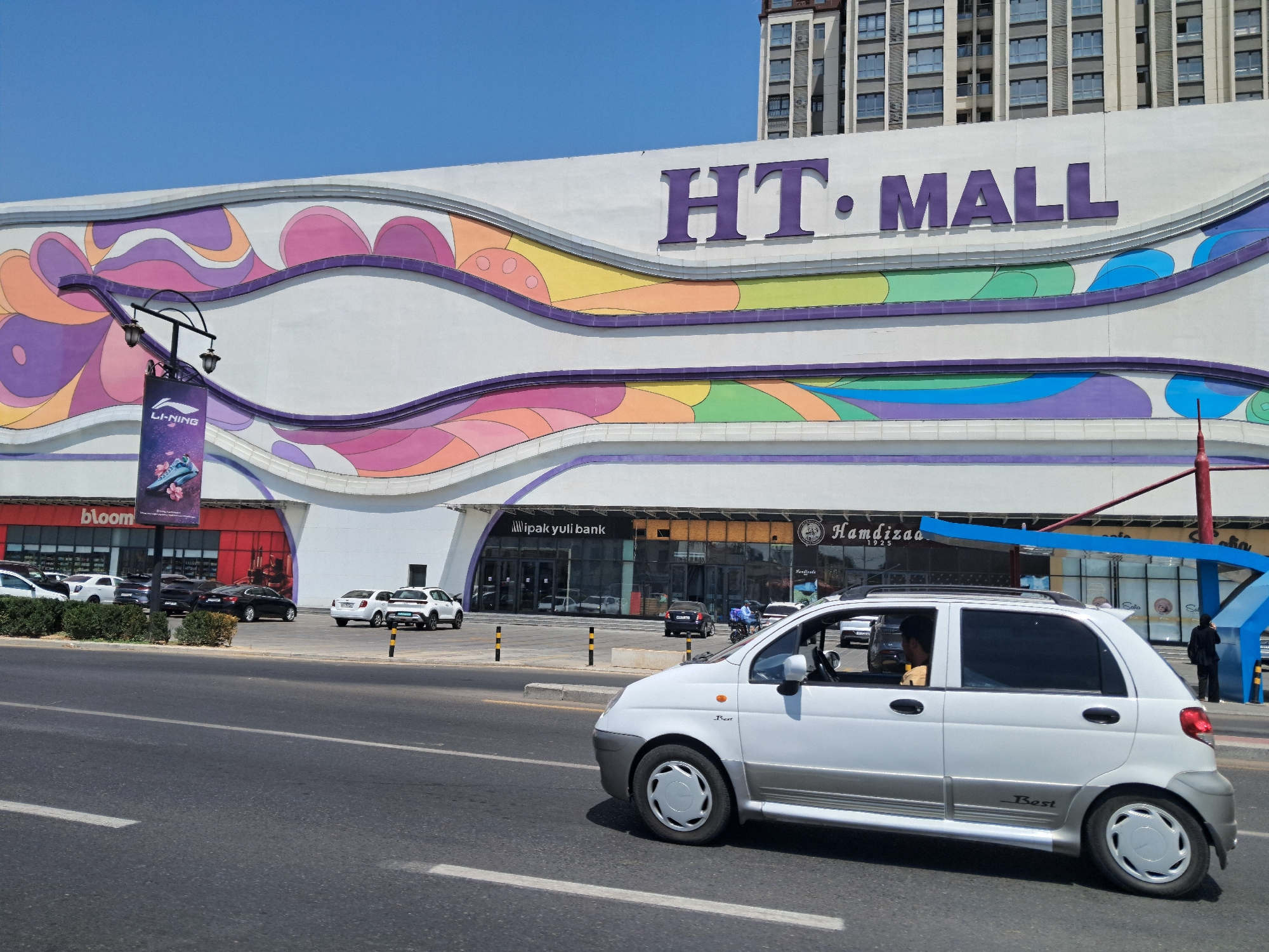 A wide shot captures the exterior of the HT Mall in Tashkent, Uzbekistan, on a clear, sunny day. The mall's facade is decorated with large, colorful, wavy patterns in shades of purple, orange, yellow, green, and blue. The prominent "HT. MALL" logo is visible in large purple letters above the entrance. In front of the mall, a row of cars is parked. The foreground features a busy street with a white car driving on the right side of the frame. The car has tinted windows and the word "Best" is visible on its side. A person can be seen driving the white car. To the right of the street, there's a blue bus stop shelter with a person standing inside. The background shows a tall apartment building. The street is asphalt with white lane markings.