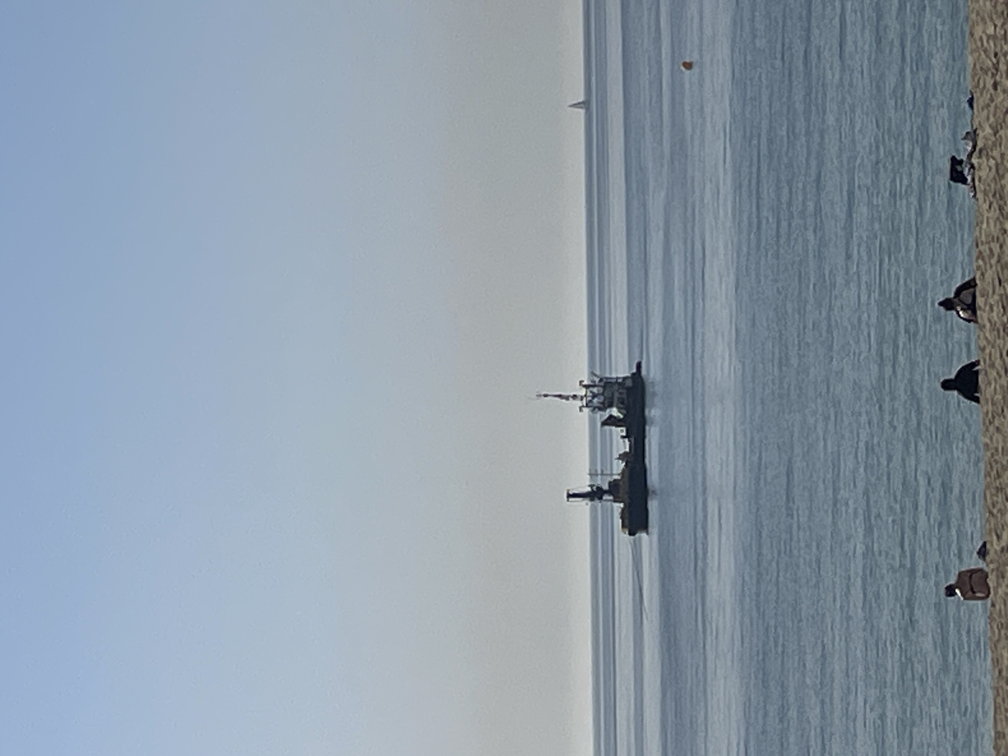 The image depicts a beach scene along the coast of Barcelona, Spain. The sky is light blue and hazy. The water is a lighter shade of blue and appears calm. Two ships are visible in the water, positioned near the center of the frame. Along the right edge of the image is a sandy beach, where several people are scattered, some sitting and others standing. In the distance, a sail is barely visible on the water.