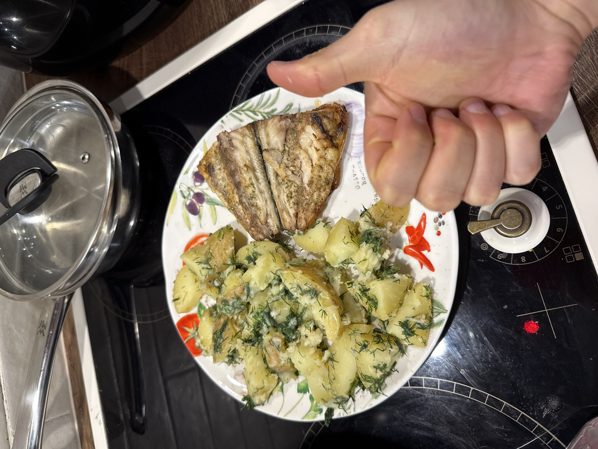 The image shows a plate of food consisting of a piece of grilled fish and boiled potatoes seasoned with dill. The plate is placed on a black stovetop next to a stainless steel pot with a lid. A human hand is partially visible, with the thumb extended. The setting appears to be a domestic kitchen. I don't have the information available to determine if this is Sofia, Bulgaria.