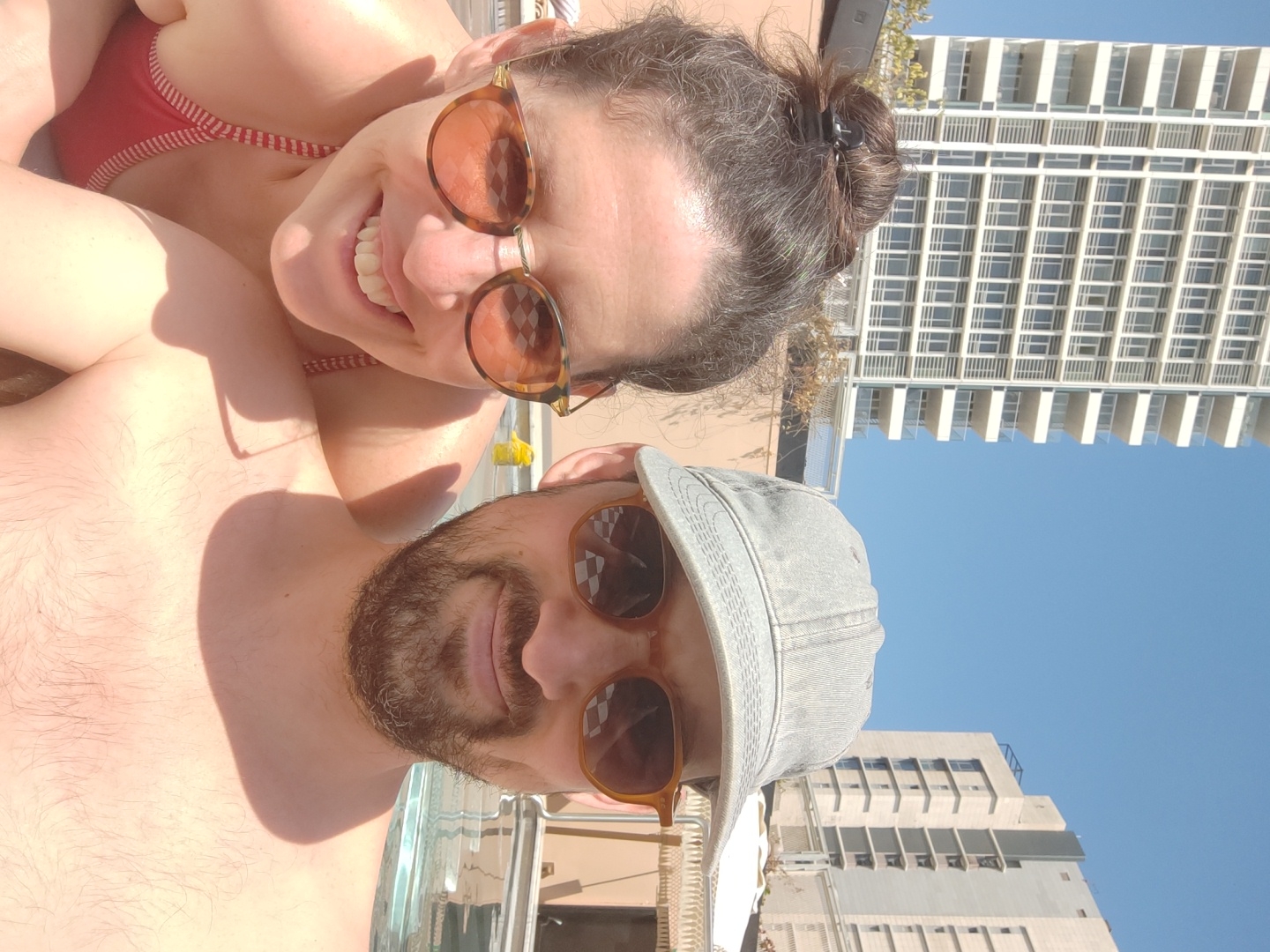 This vibrant image is a selfie of two individuals, a woman and a man, captured outdoors on a sunny day. The woman, prominently in the foreground, smiles broadly, showing her teeth, and wears a red and white striped bikini top. She has dark hair tied back in a bun with a clip and wears tortoise-shell framed sunglasses that reflect a checkered pattern.
Partially visible below her is a man, also wearing tortoise-shell framed sunglasses and a light-colored, possibly denim, baseball cap. He has a beard, and his bare chest is visible, indicating warm weather. Both individuals appear relaxed and happy, with the woman's wide smile conveying joy.
The background features a clear, bright blue sky and a distinctive urban skyline, consistent with Tel-Aviv, Israel. Several modern high-rise buildings are visible, most notably a tall white building with numerous windows and balconies on the right, and a shorter, darker building on the left. The strong sunlight casts distinct shadows, suggesting it is midday or early afternoon. The scene conveys a leisurely moment, possibly on a rooftop or balcony overlooking the city. There is no visible text in the image.