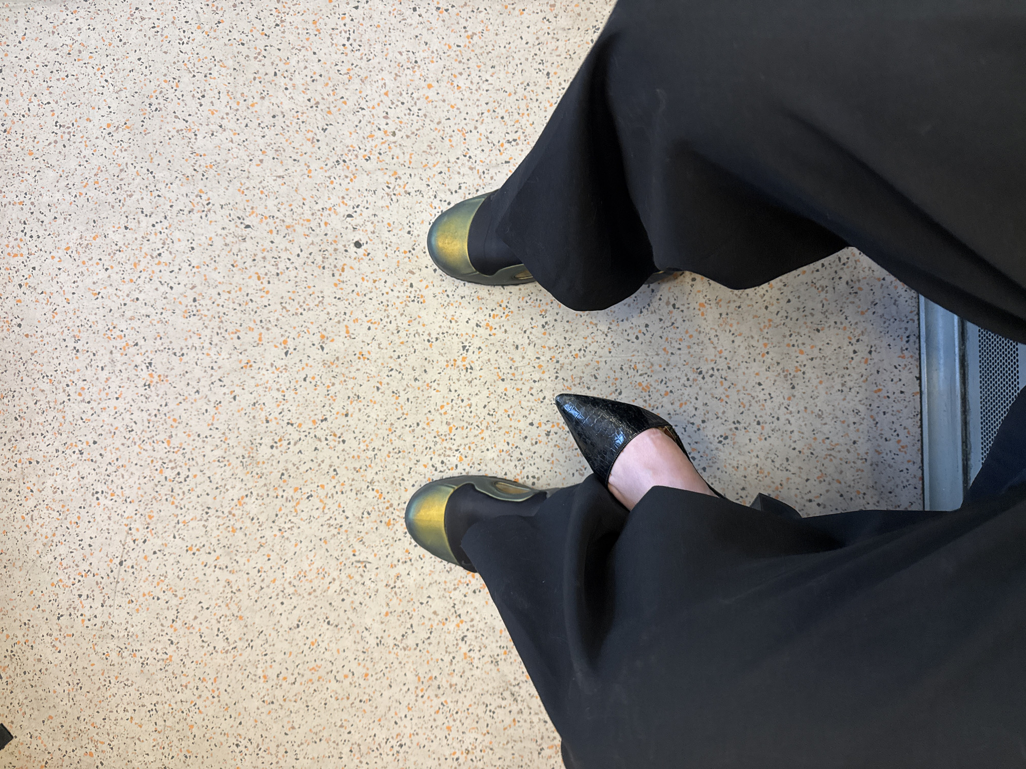 An overhead shot captures the lower half of a person seated indoors, likely in a public space in London, United Kingdom. The individual wears wide-leg black trousers, which fall loosely, partially obscuring their feet.
The footwear is a notable detail, with two distinct styles. The left foot is shod in a closed-toe, flat or low-heeled shoe that exhibits an iridescent, possibly metallic, finish, shimmering with hues of gold, green, and blue. In contrast, the right foot wears a black, pointed-toe pump with a textured surface, suggestive of crocodile or snake skin. A small sliver of bare skin is visible between the right shoe and the trouser hem.
The floor beneath is a light-colored, speckled surface, resembling terrazzo, embedded with numerous small flecks in shades of orange, grey, and black. Along the far right edge of the frame, a glimpse of a grey metallic structure with a perforated or mesh pattern is visible, hinting at a public seating arrangement, perhaps within a waiting room, train station, or airport. The person appears to be in a relaxed, seated position, indicative of waiting. No specific time of day, weather conditions, or visible text can be discerned from the image.