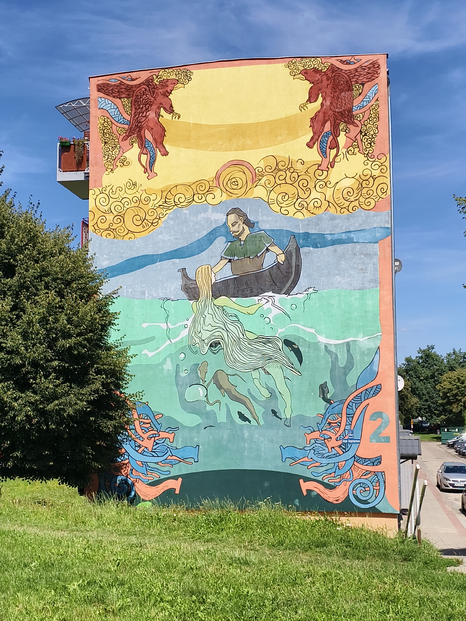 The image shows a large, colorful mural on the side of a building in Tczew, Poland. The mural depicts a scene with mythological or fantasy elements.
The top section of the mural features two winged, dragon-like creatures with red bodies and brown wings against a yellow and orange sky. Swirling patterns in yellow and blue fill the space between them.
Below this, a man with long brown hair and a beard, wearing a green tunic, is rowing a dark canoe. He is looking towards the right. In front of the canoe, a long-haired, ethereal figure with flowing light green hair and a light green body appears to be emerging from the water. Several small, dark fish are swimming around this figure. The water is depicted in shades of light blue and green with white wavy lines and circles.
The bottom section of the mural includes abstract, organic shapes in blue and orange, resembling tentacles or intertwined limbs. The number "2" in orange is prominently displayed on the right side of this section. A small plaque with Polish text is visible beneath the "2".
The building itself is painted with this mural. A balcony with plants is visible on the left side of the building, extending above the mural. The building is situated in an urban environment, with a grassy area in the foreground and trees and a street with parked cars visible in the background. The sky is clear and blue with some wispy clouds. The sun is shining, casting shadows.