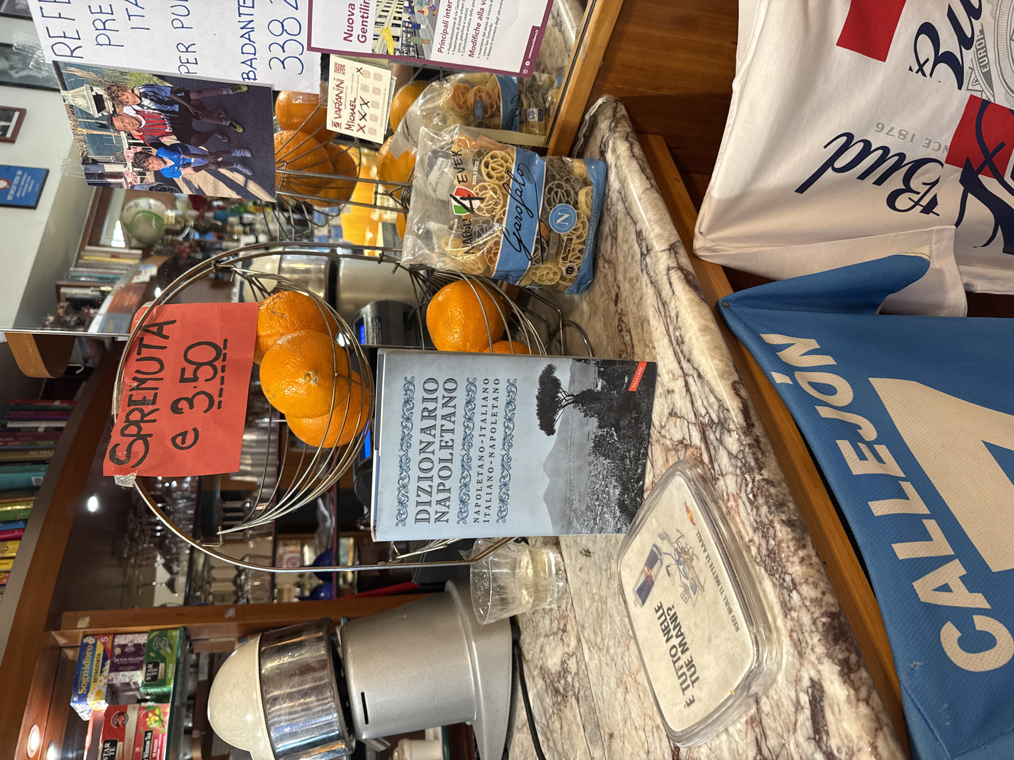Here's a description of the image:
The image depicts a counter or shelf in what appears to be a shop or cafe, likely located in Italy. The scene includes several items: oranges displayed in a metal basket with a handwritten sign indicating "SPREMUTA" (fresh juice) and a price of 3.50, a bag of Garofalo pasta, and a book titled "DIZIONARIO NAPOLETANO" (Neapolitan Dictionary). Also visible are various products, including teas and a small container with a Red Bull advertisement. The background features a variety of items on shelves, suggesting a retail environment. A Budweiser promotional shirt and a blue flag with the word "CALLEJON" are present. Based on the Gentilin ad, this location is potentially in the city of Milan, Italy.