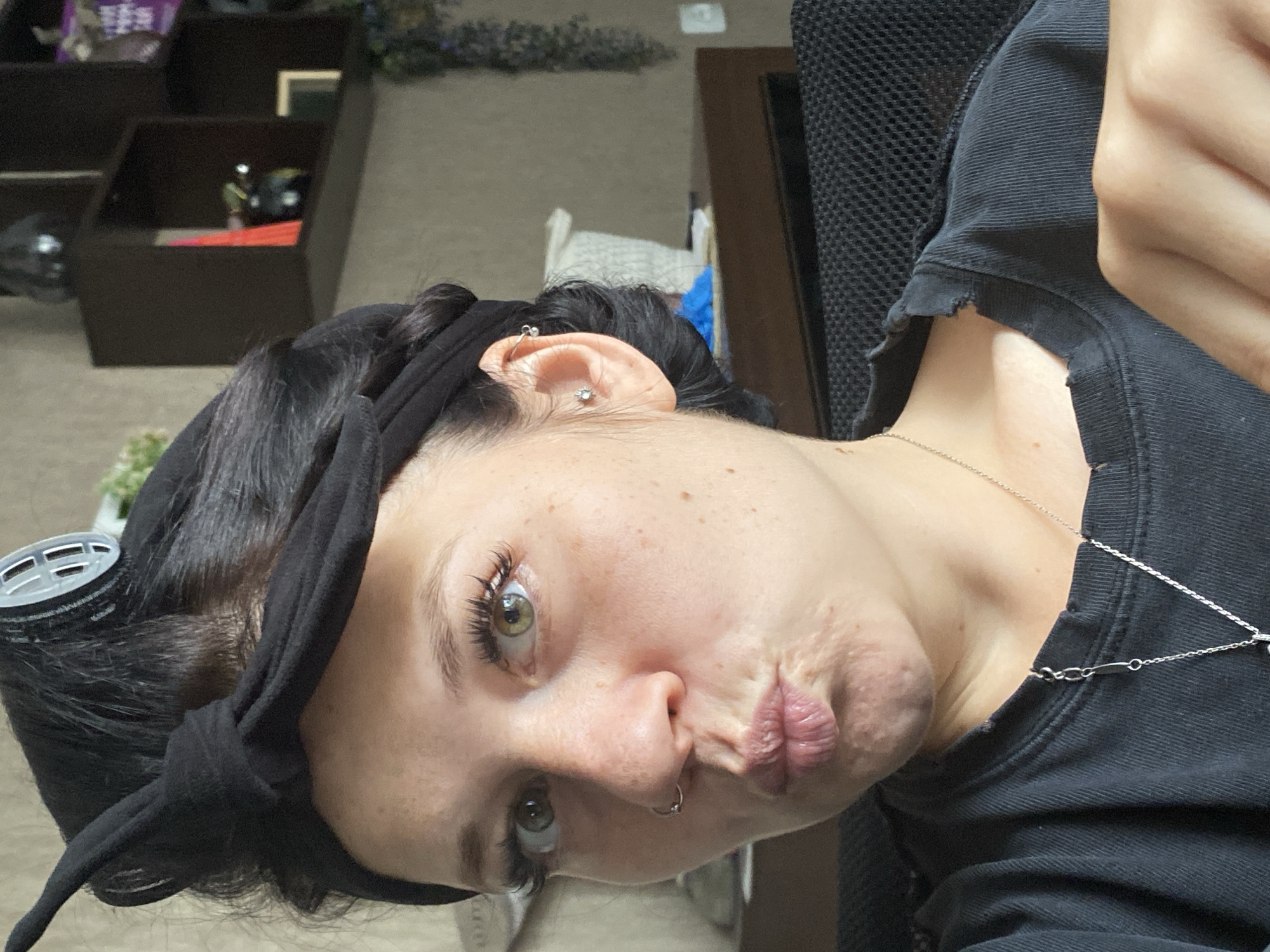 The image shows a close-up, medium shot of a woman with her hair in rollers and a black headband, making a duck face. She has a nose ring and a second piercing in her left ear with a stud. She is wearing a black t-shirt and a silver necklace. The background appears to be an indoor setting, possibly a room in a home or an office, with shelves and plants visible. The lighting suggests it is daytime. There is no indication of the specific location being in Chaikovsky, Russia.