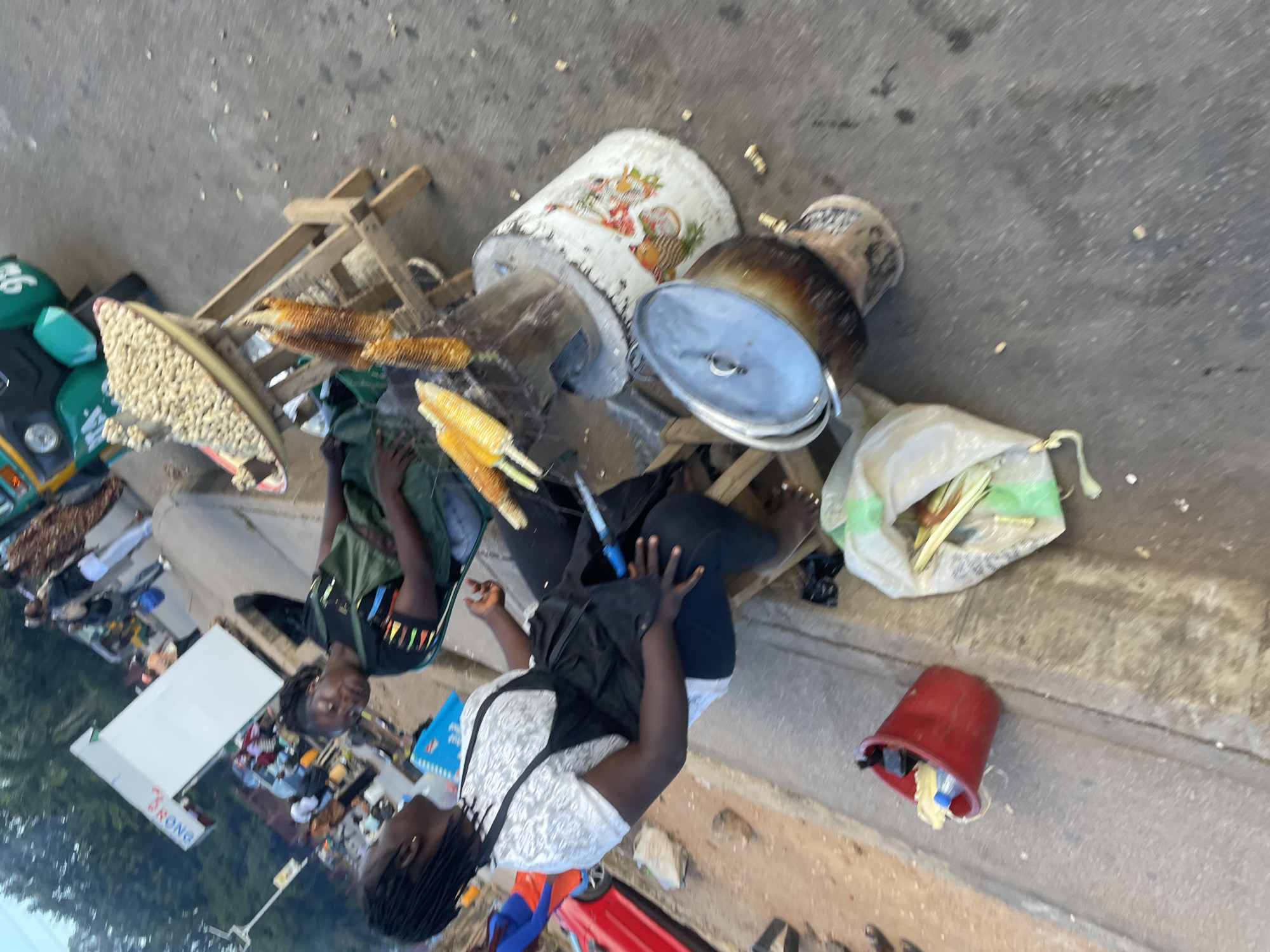 In Jos, Nigeria, a street vendor is seen preparing food. In the foreground, a woman wearing an apron over a white patterned shirt and dark pants is seated. To her right, another woman, also seated and wearing a dark apron over a black t-shirt, appears to be sleeping. Behind them, a wooden stand holds a large bowl filled with nuts, and several ears of corn are being grilled over an open fire. A large pot with a lid sits on a makeshift stove. To the left of the vendors, a red bucket is filled with various items, and a large bag containing what appears to be corn husks lies on the ground. In the background, several other individuals are visible, along with a green motorized tricycle and a white rectangular structure. The scene depicts a busy street market environment.