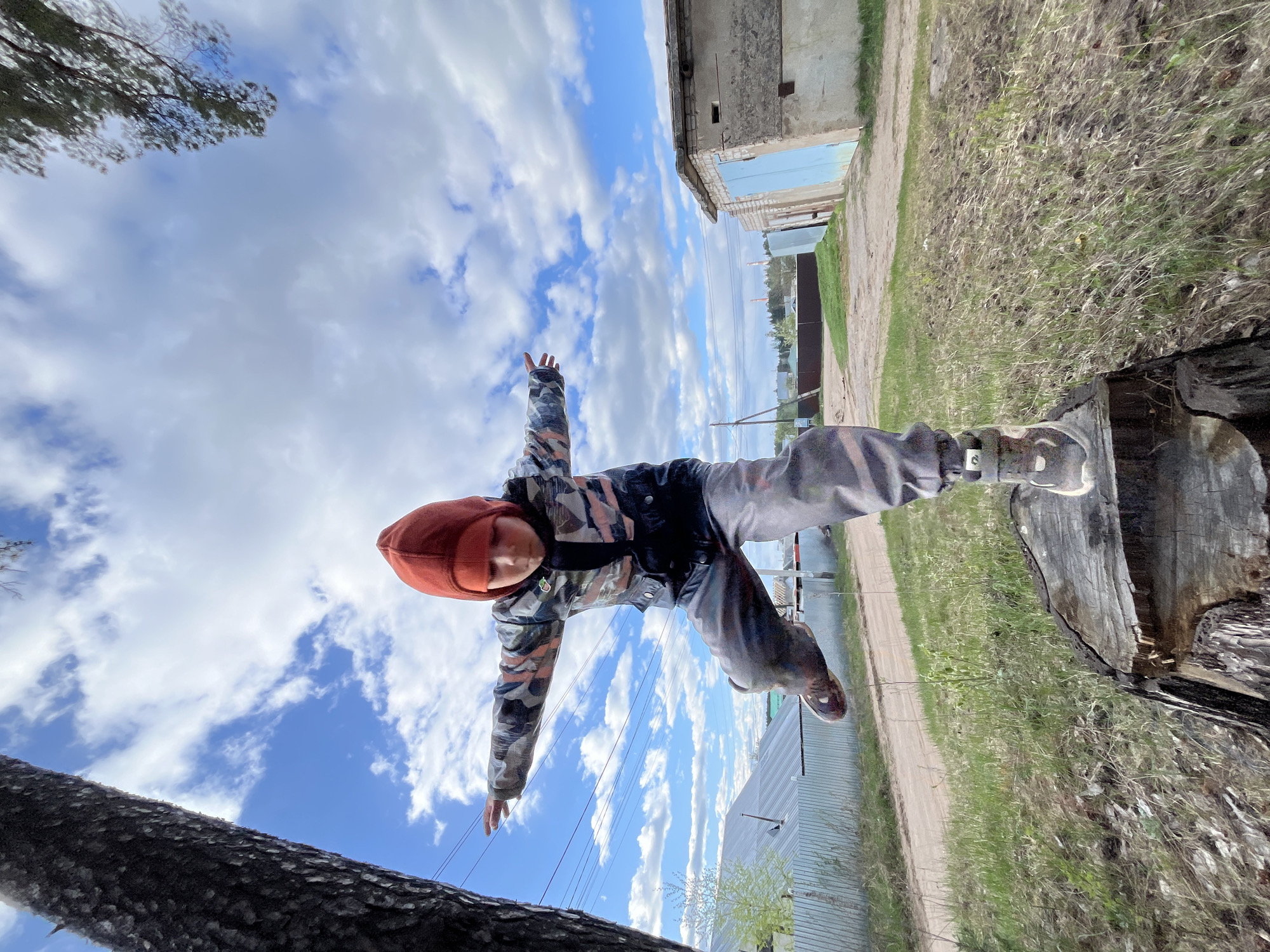 A child in camouflage clothing and a red beanie stands balanced on a weathered tree stump in an outdoor area. The child's arms are outstretched, suggesting a sense of balance or play. The background includes a building with blue accents, a grassy area, a dirt path, and a sky filled with clouds and patches of blue. The photograph appears to be taken from a low angle, looking upwards. While the specific location in the city of Chaikovsky, Russia is not identifiable from the image alone, the setting appears to be a somewhat rural or suburban area within the city.