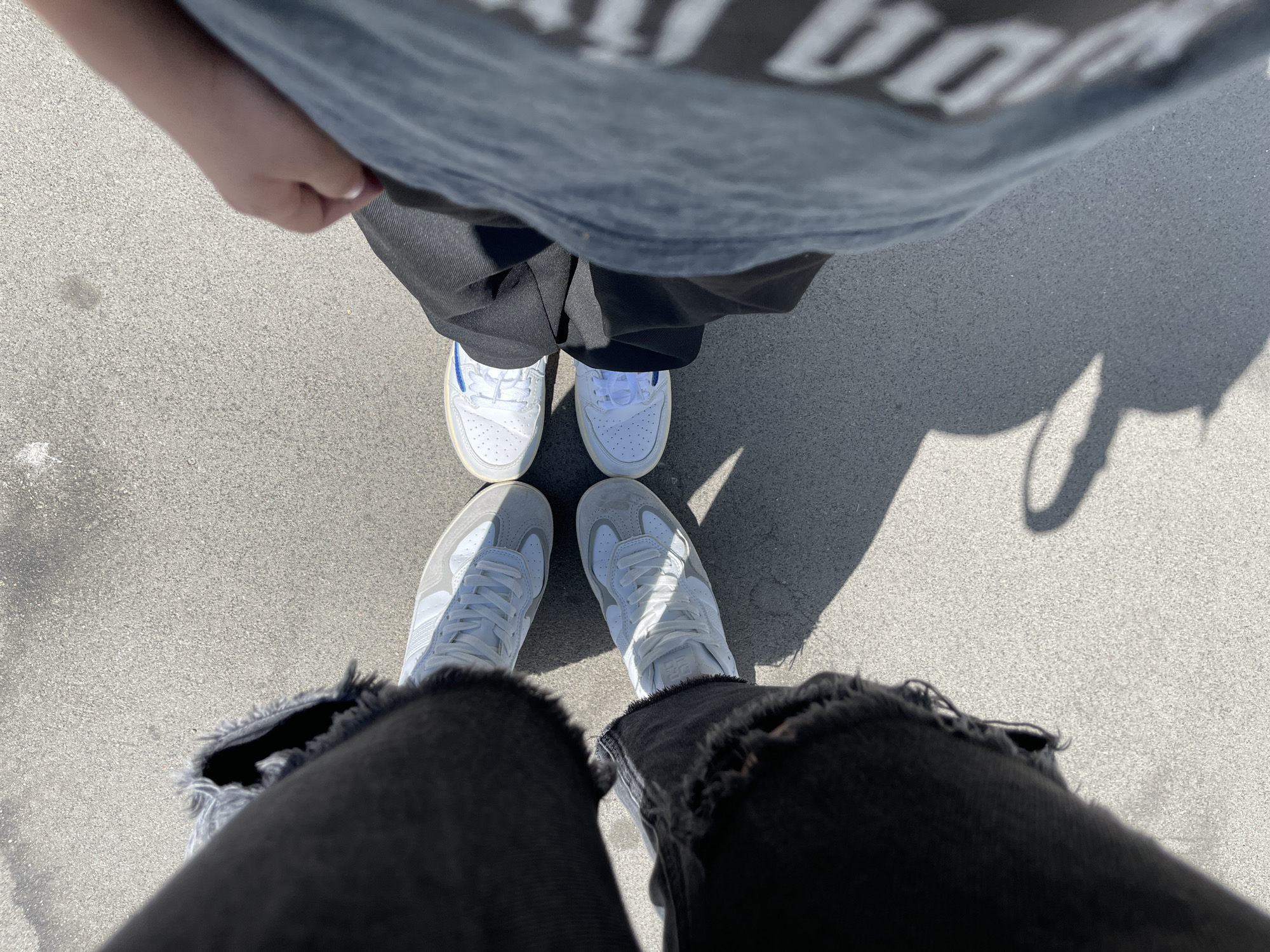 An overhead, high-angle shot captures two individuals standing on light grey asphalt or concrete pavement under bright, direct sunlight. The scene is likely outdoors, possibly in Moscow, Russia, as provided context.
In the background, one person is seen from the chest down, wearing a grey t-shirt with blurry white lettering visible across the chest area. They are also wearing dark grey or black trousers and white low-top sneakers featuring distinctive blue stripes or accents on the sides. Their hands are slightly visible near their waist.
In the foreground, the second individual's legs and feet are visible, wearing black distressed jeans with frayed hems and a pair of white and light grey low-top sneakers. Both individuals are positioned facing each other, with their feet close together.
The clear, sharp shadows cast on the ground by both people indicate strong sunlight, suggesting it is likely midday or early afternoon. The pavement has a granular texture. No specific activity beyond standing is discernible.