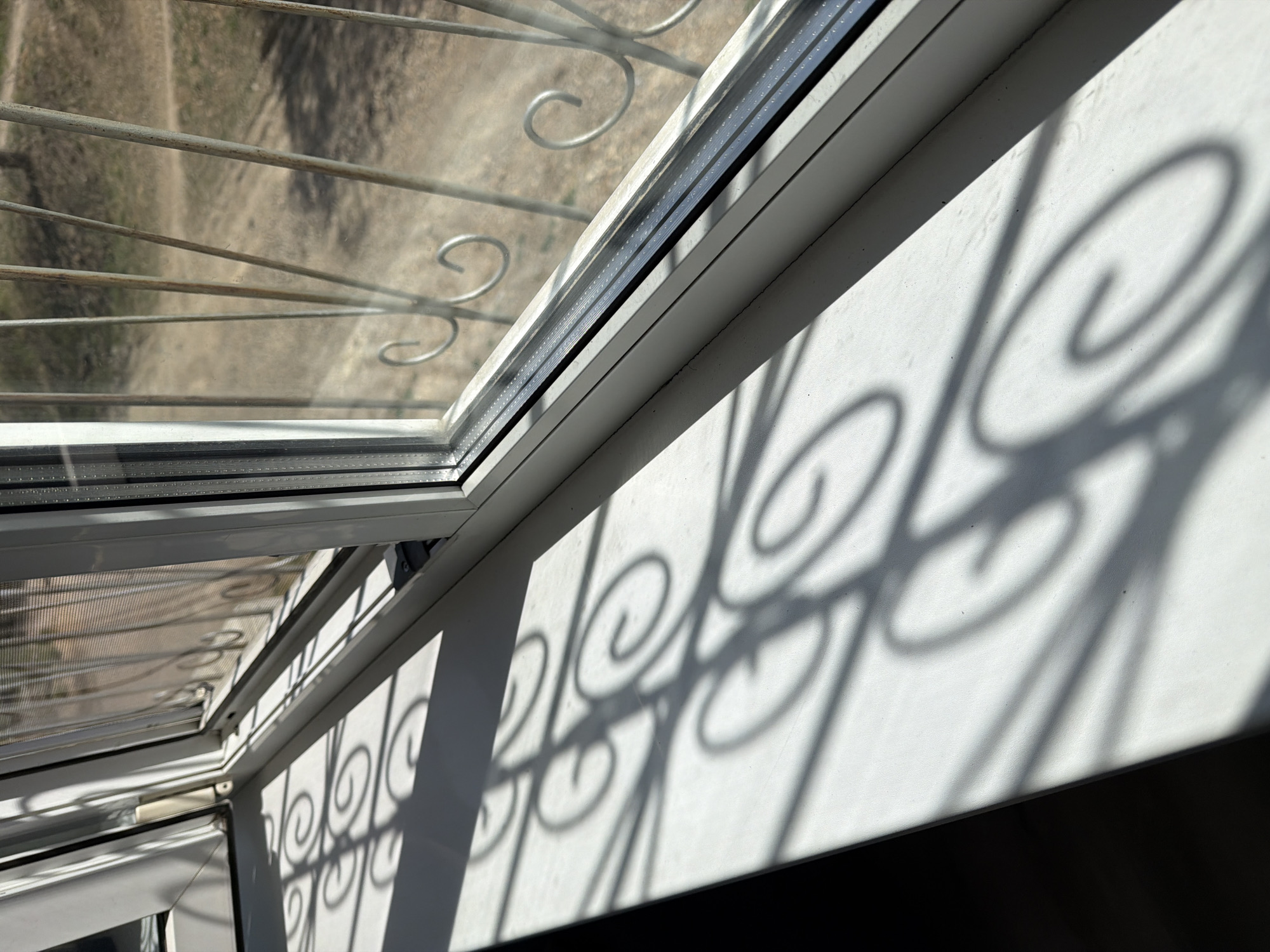The image shows an interior view of a window with a decorative iron grill. The window frame is white, and the shadows cast by the grill create an intricate pattern on the adjacent white wall. The view through the window appears to be a landscape of dry earth or a field. The location of the scene is in the city of Birobidzhan, Russia.