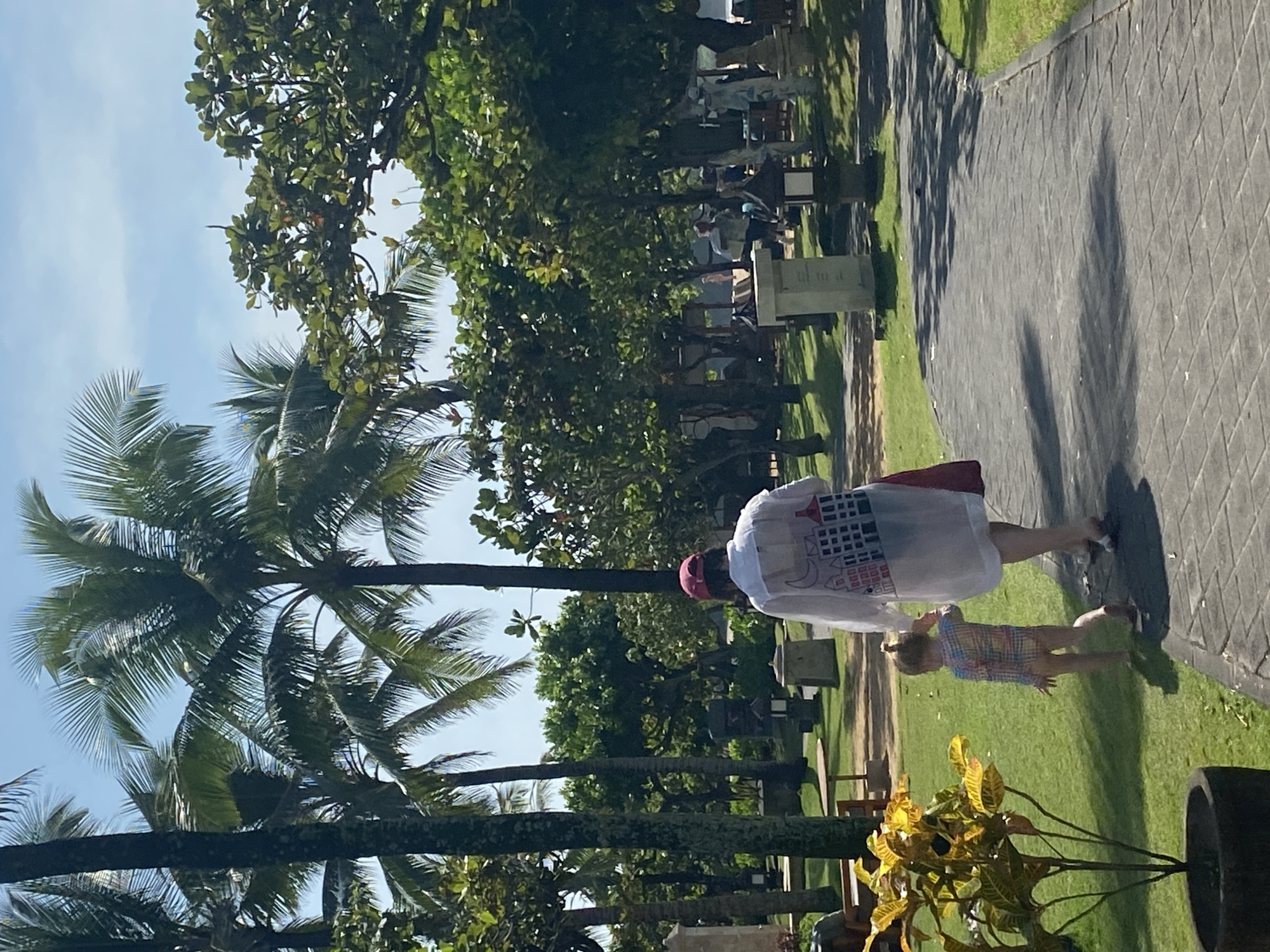 The image shows a daytime scene in Nusa Dua, Indonesia. A paved pathway is bordered by green grass and tropical vegetation, including palm trees. A woman in a white shirt with a cityscape design on the back and a pink hat is walking alongside a young child. There are other people and park benches in the background. The sky is blue.