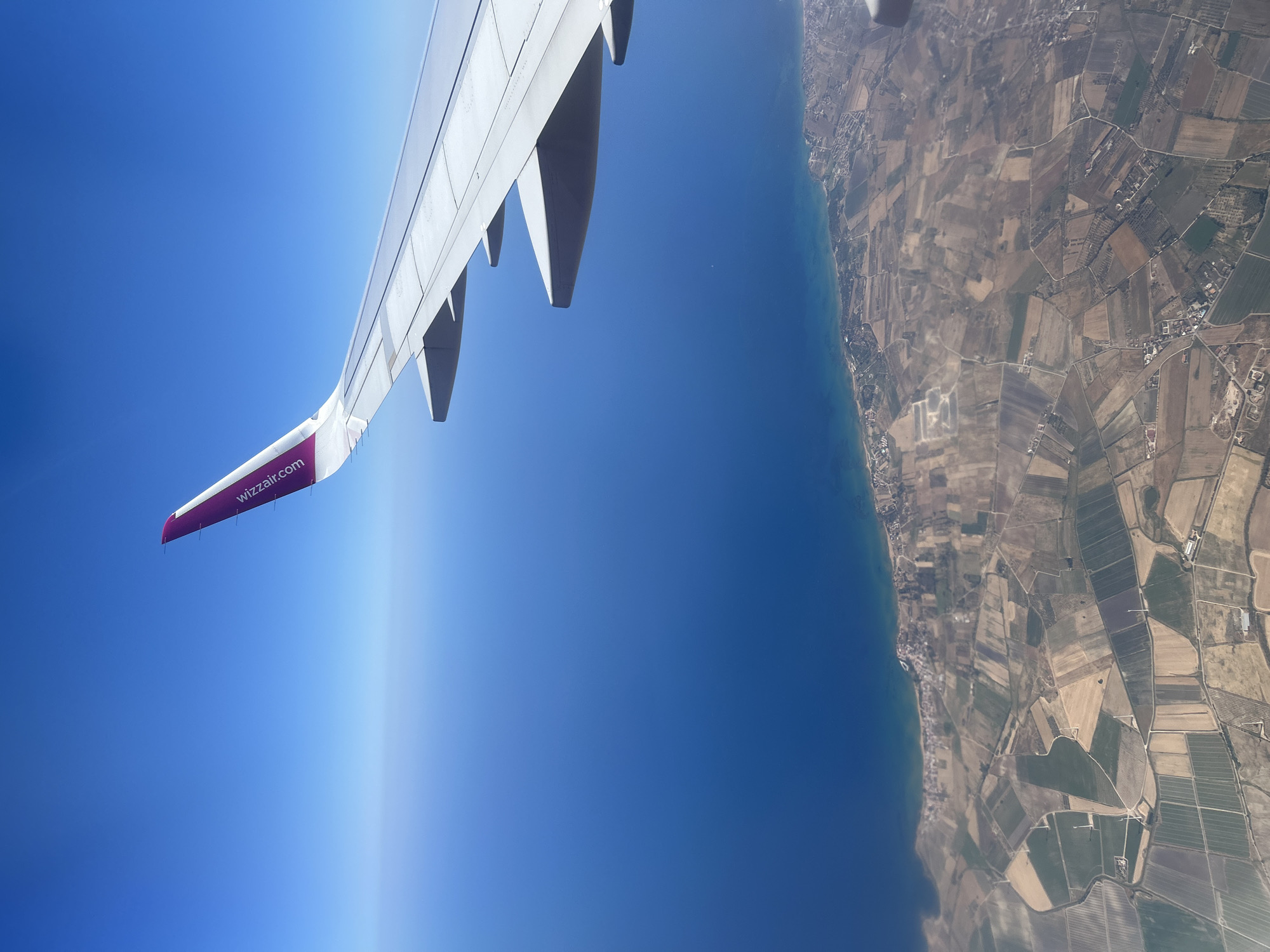 The image captures an aerial view from an airplane window, looking downwards. A prominent airplane wing, partially white and partially a deep purple with the text "wizzair.com" visible, occupies the left side of the frame. Below the wing, the deep blue expanse of the sea stretches out, meeting a coastline. The land adjacent to the sea is a patchwork of agricultural fields, divided by roads and featuring scattered buildings and structures, indicative of rural or semi-rural settlement.
The scene suggests the airplane is in flight, likely during daylight hours due to the bright illumination. The sky above is a clear, bright blue, transitioning to a lighter shade near the horizon, hinting at a sunny day. The curvature of the Earth is subtly suggested by the gradient of the sky. The location context provided is Lamezia Terme, Italy, which aligns with the coastal and agricultural landscape visible. There is no discernible activity or event taking place, other than the flight itself. The image evokes a sense of travel and the vastness of the view from above.