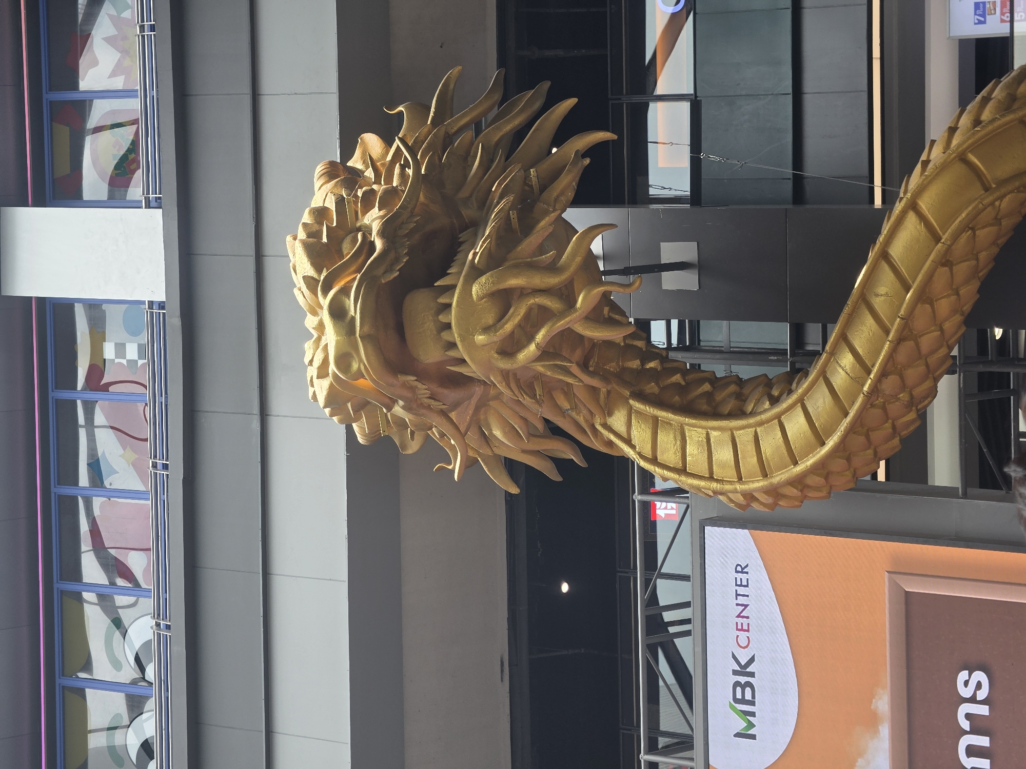A golden dragon statue adorns the MBK Center in Bangkok, Thailand. The statue, appearing to be made of metal, is positioned in front of the building, with the building's architecture and signage clearly visible in the background. The "MBK Center" logo is prominently displayed, along with what appear to be Thai characters. The angle of the photograph is vertical. The building itself includes stained glass windows.