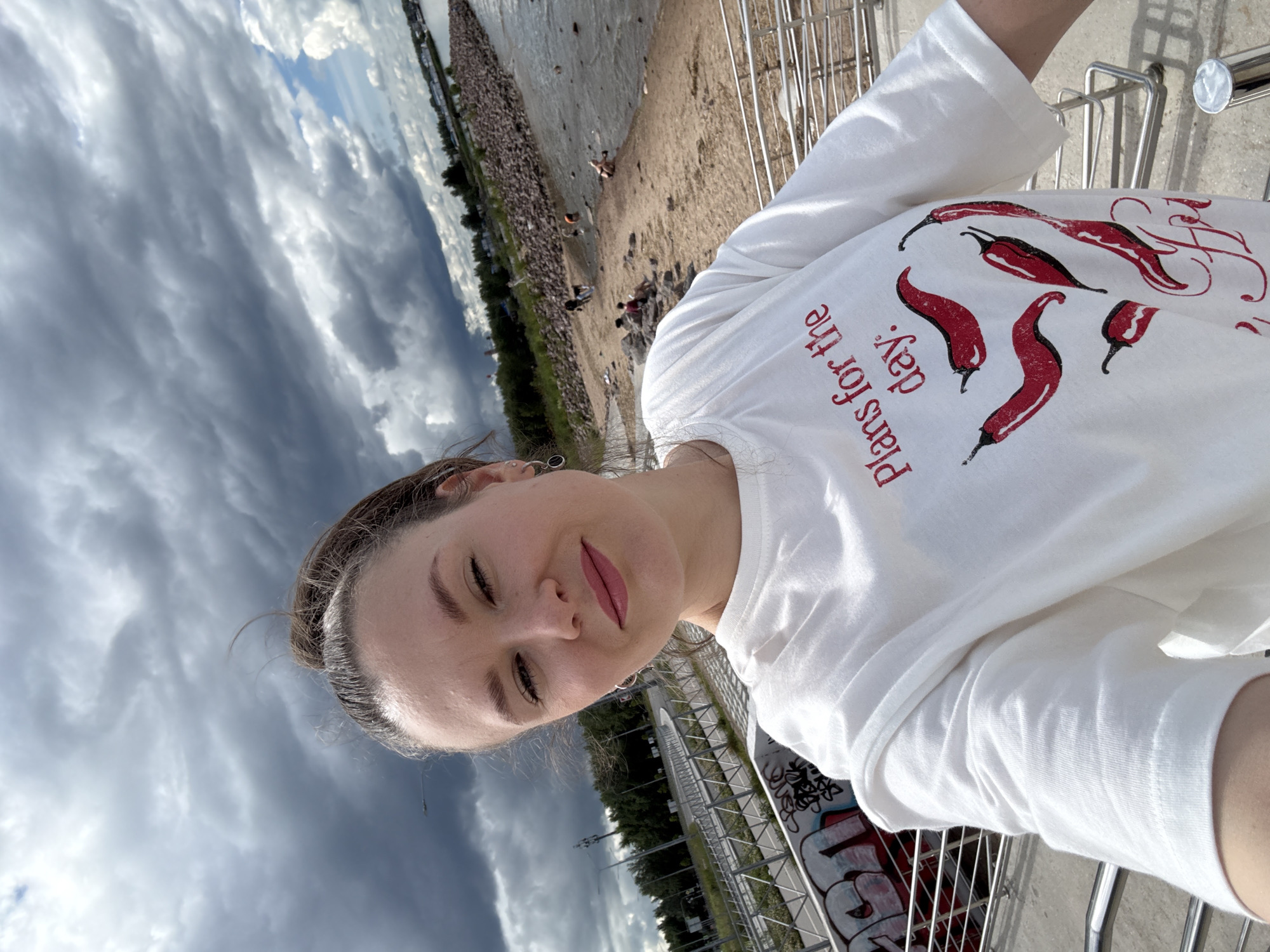 A young woman with her hair pulled back takes a selfie with a dramatic, cloudy sky as the backdrop. She is wearing a white long-sleeved t-shirt with a graphic print of red chili peppers and the words "Plans for the day:". Her eyes are closed and she is smiling, wearing lip gloss. She is leaning against a metal railing.
In the background, a body of water stretches out to the horizon, bordered by a rocky shore and a sandy beach. Several people are scattered on the beach, some appearing to be sitting or lying down, while others are walking. A line of trees and some buildings can be seen further inland. The weather appears overcast with heavy, dark clouds suggesting an impending storm. The scene is set outdoors, likely near a coastline or large body of water. Graffiti can be seen on a wall in the lower right corner of the image. The overall mood is one of casual outdoor activity under a moody sky.