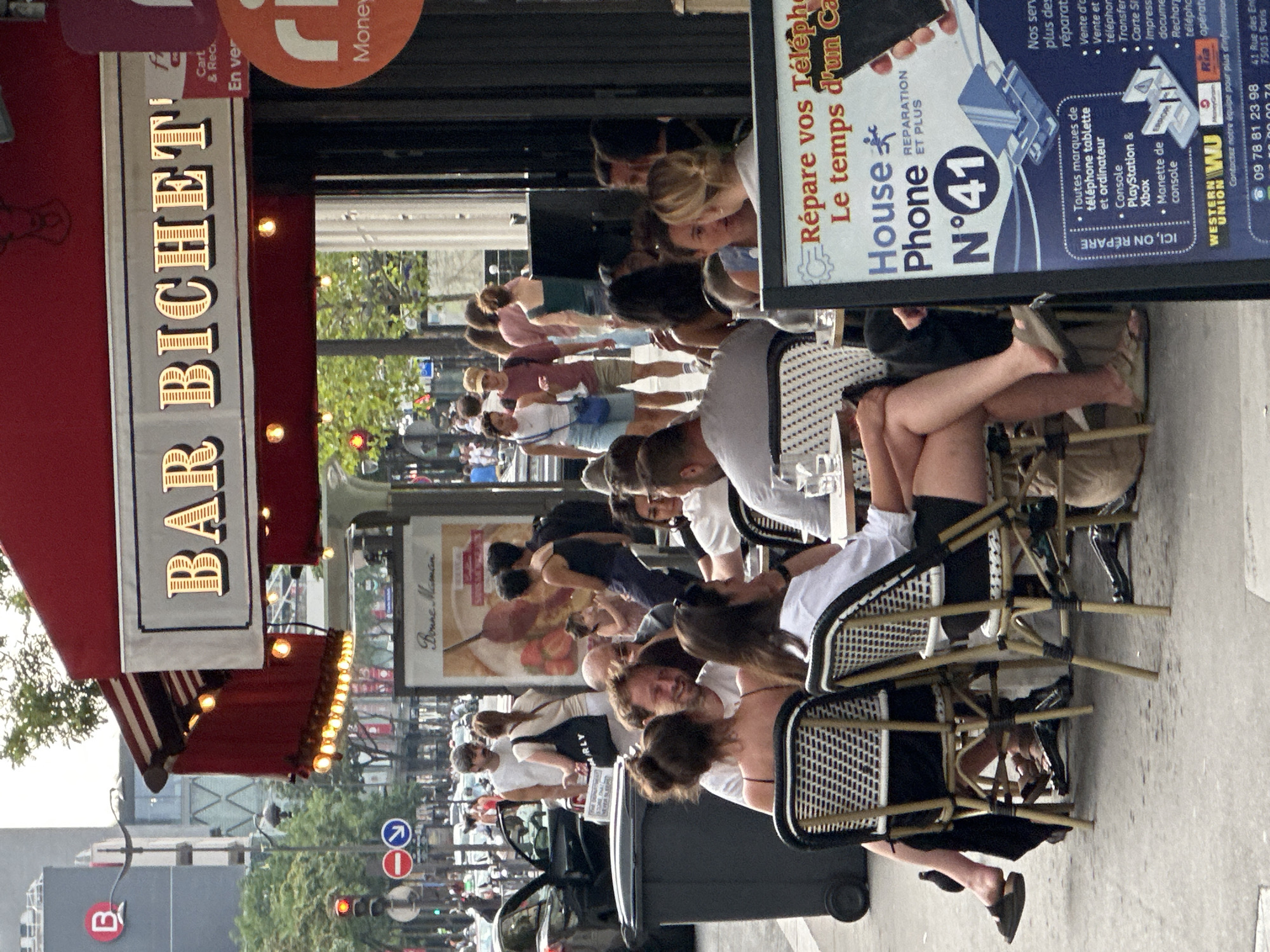 This image depicts an outdoor seating area of a Parisian cafe, specifically "Bar Bichet," located in Paris, France. The cafe has a red awning and illuminated signage. Several patrons are seated at tables on the sidewalk, engaged in various activities such as dining and conversing. One table in the foreground features a group of people, with a woman wearing a white shirt and black shorts in the center. To the right, a sign with French text advertises phone repair services. In the background, a street scene unfolds with pedestrians, vehicles, and buildings. The overall atmosphere is that of a bustling urban environment.