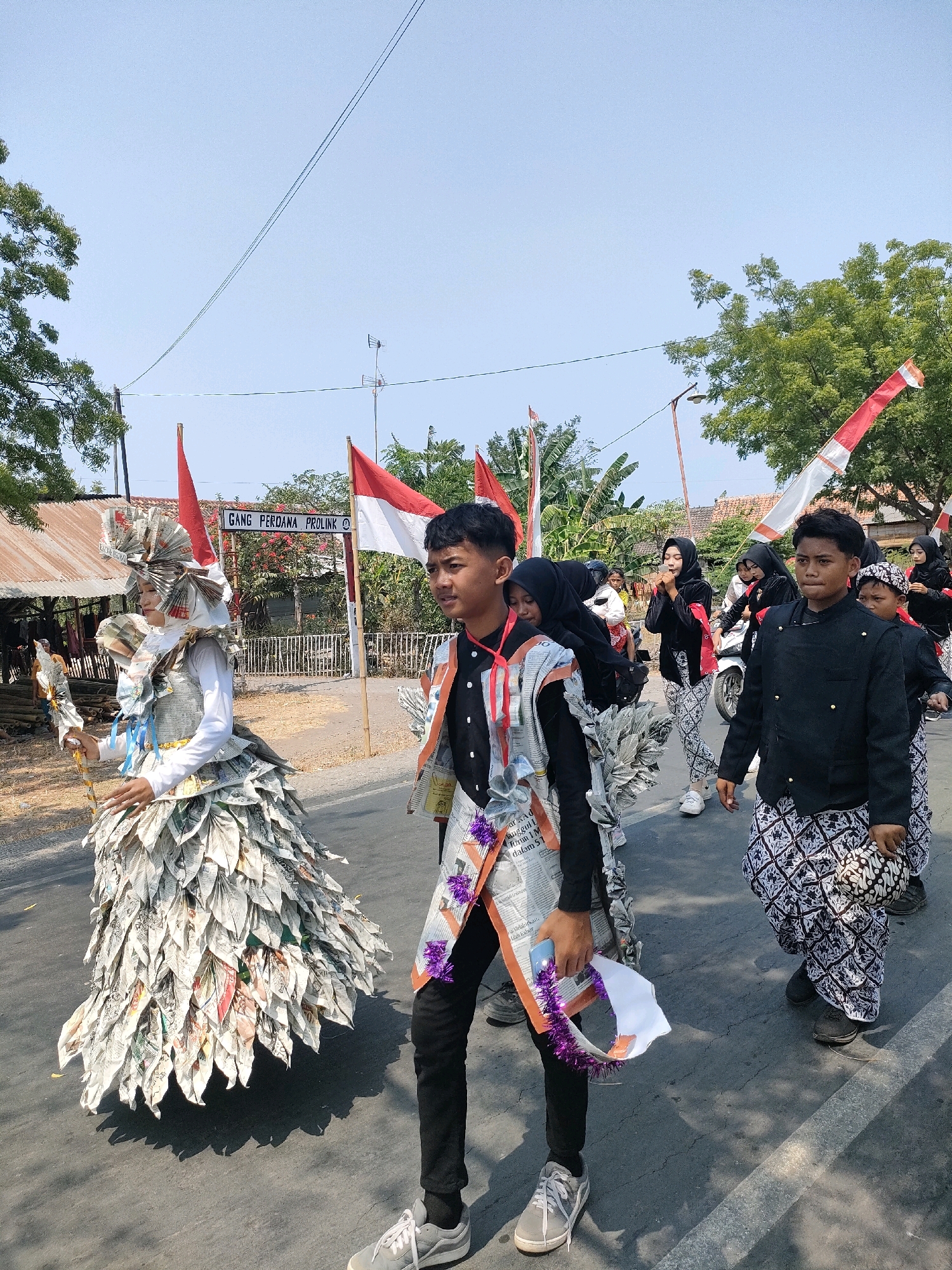 In Tongaswetan, Indonesia, a daytime parade is underway on a paved road under a clear sky. The parade features individuals in traditional attire and elaborate costumes. A central figure wears a voluminous dress constructed from layered, folded newspaper, resembling petals, with a matching headpiece and accessories. Beside them walks a young man in a black jacket and patterned trousers, holding a small white flag adorned with purple tassels. Other participants, including young women in headscarves and traditional clothing, follow, some on foot and one on a scooter. Indonesian flags, red and white, are prominently displayed throughout the scene. The background includes a sign reading "GANG PERDANA PROLINK," a rustic building with a corrugated metal roof, trees, and a light pole. The atmosphere suggests a community celebration.