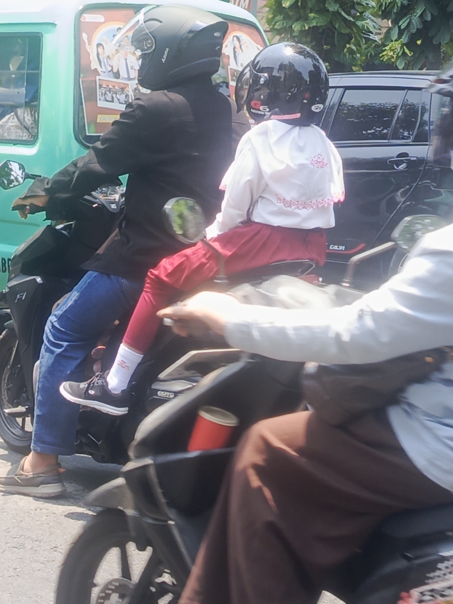 This is a close-up, eye-level, medium shot of two individuals riding a motorcycle on an urban street in Surabaya, Indonesia. The foreground shows the leg and torso of the rear passenger and the right leg of the driver. The driver, wearing blue jeans and black sneakers, is operating the motorcycle. The rear passenger, a child, is wearing a white top with pink floral embellishments and red pants, with white socks visible. Both individuals are wearing black helmets. A red cylindrical object, possibly a can, is attached to the motorcycle's frame. In the background, a green vehicle is partially visible on the left, and a dark car is on the right. The environment suggests a sunny day with some foliage visible behind the car.
