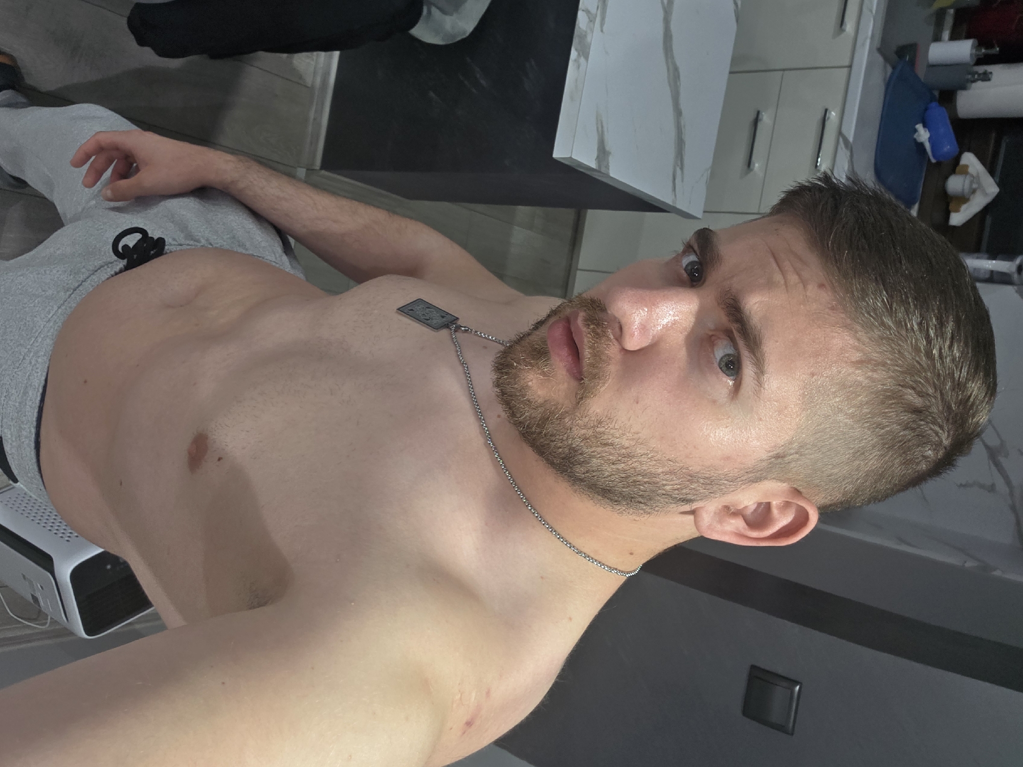 The image depicts a shirtless man with a beard and short brown hair wearing grey sweatpants and a silver chain necklace. He is indoors, with a countertop visible behind him, possibly in a kitchen or bathroom based on the countertop material and visible cabinets. Objects such as a blue bottle and rolls of white material are partially visible in the background. A white, rectangular object with ventilation slots is near the bottom left of the frame. The man's expression is neutral, looking directly at the camera. There is no information indicating that the location is Yagodovo, Bulgaria.