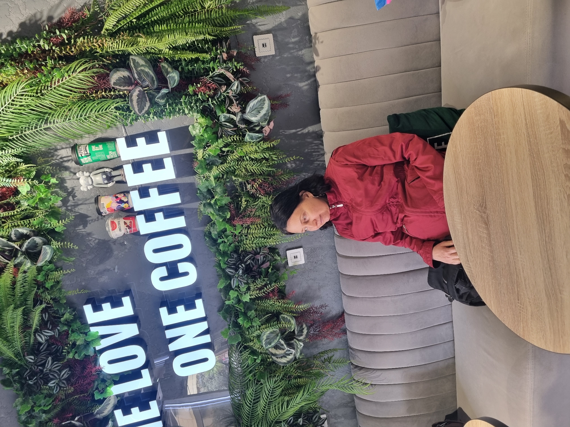The image captures a cafe interior, likely in Sochi, Russia, given the text on the signage. A woman with dark hair, wearing a red jacket, is seated on a gray, segmented couch. Next to her is a round, light-brown table. Behind the couch and woman is a wall adorned with abundant greenery, encompassing various ferns and leafy plants. Inset into this decorative wall is the phrase "I LOVE ONE COFFEE" in large, illuminated, white block letters. Several small objects and cans of coffee adorn the wall, nestled within the greenery. The lighting appears to be soft and ambient, suggesting an inviting atmosphere.