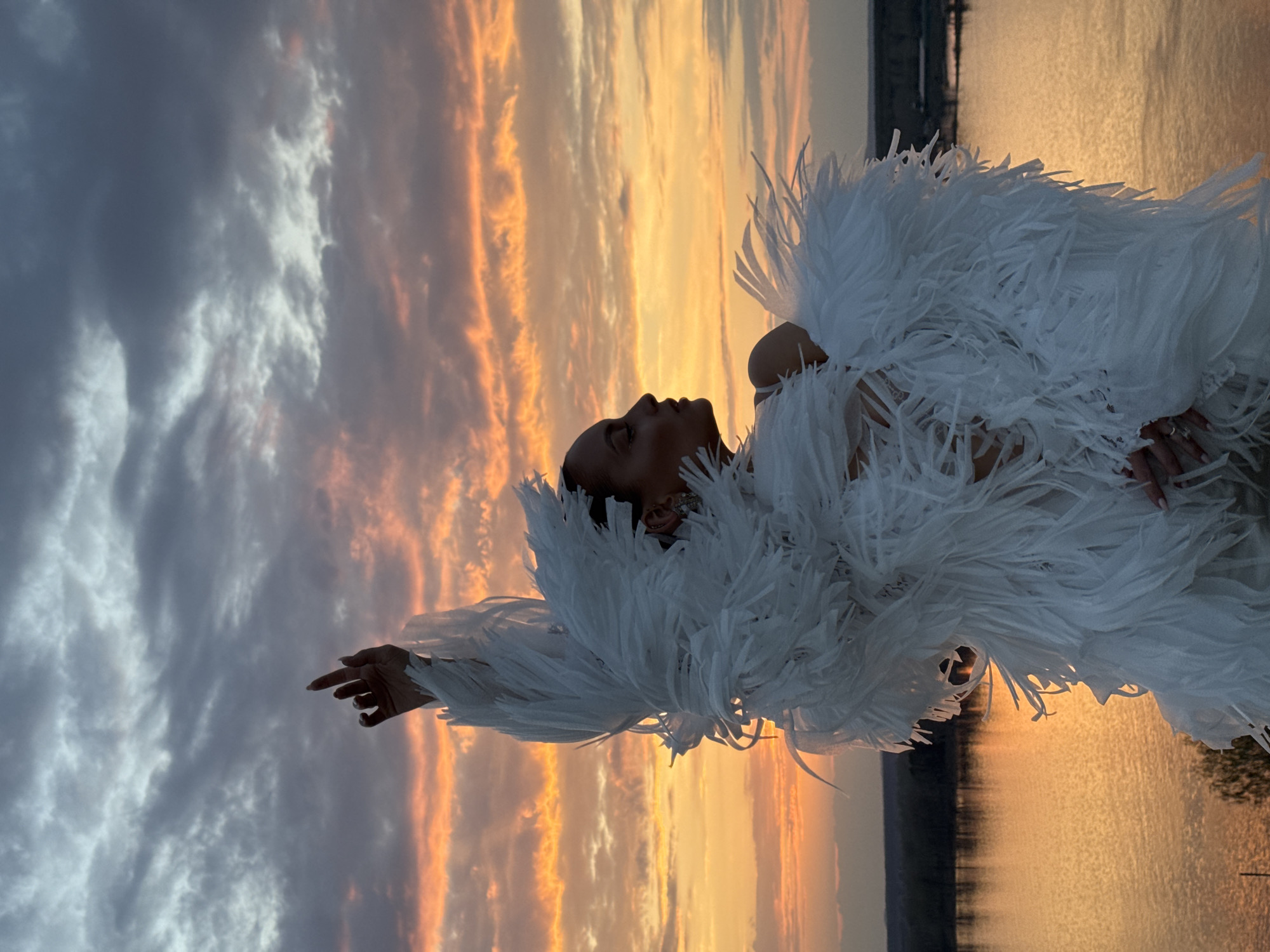 A vertical, full-shot image shows a person from the side, facing upwards, in a white feathered garment with outstretched arms, set against a sunset sky and a body of water. The sky is a mix of gray, white, and orange hues, suggesting either dawn or dusk. The body of water appears calm, reflecting the colors of the sky. The person's profile is visible, and they appear to be of African descent. They are wearing jewelry on their ears and fingers. The feathered garment is large and fluffy, obscuring most of the person's body. In the background, there is a darker line suggesting the horizon, possibly distant land. The image appears to be taken outdoors, likely at a scenic location. There is no explicit indication of the image's location in Ольховка, Russia.
