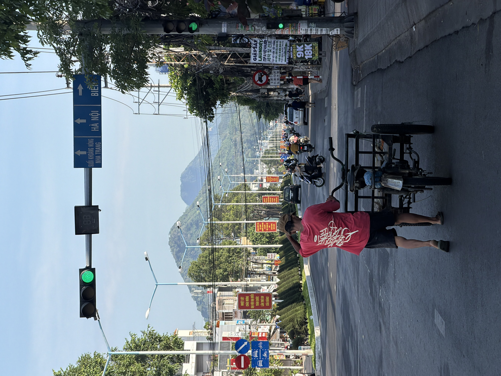 The image shows a street scene in Bac Nha Trang Ward, Vietnam, with a man pushing a three-wheeled cart. The man is wearing a red t-shirt and dark shorts, and he is walking away from the camera. The cart is made of metal and has a black frame. In the background, there are mountains, buildings, and traffic, including motorcycles and cars. The sky is blue with some clouds. The street appears to be paved and wet, suggesting it may have recently rained. Traffic lights and directional signs are visible, indicating this is an urban area with traffic control. There are also several street lamps lining the road. Advertisements and signs are posted on utility poles and buildings.