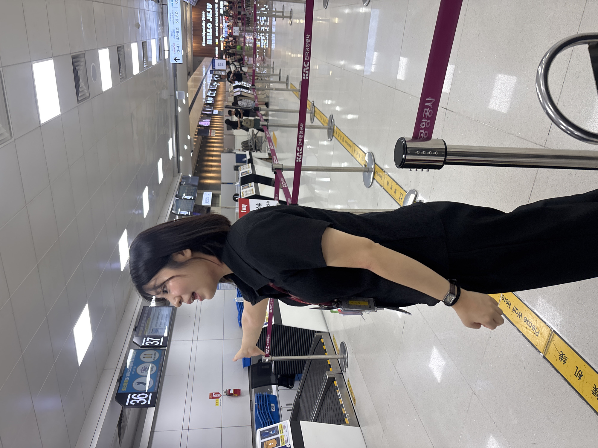 The image shows a woman standing in an airport, likely at a check-in counter. She is wearing a black shirt and has dark hair. The airport appears to be indoors, with bright fluorescent lighting. There are rows of stanchions with purple and yellow tape, indicating a queueing area. Signs in Korean and English are visible, including "KAC 한국공항공사" (KAC Korea Airports Corporation) and "Please Wait Here." Other signs indicate gate numbers and destinations like "HELLO JEJU." The woman is looking to her right and appears to be smiling or mid-sentence. The overall impression is of a typical airport scene, possibly during the day, with passengers waiting for their flights.