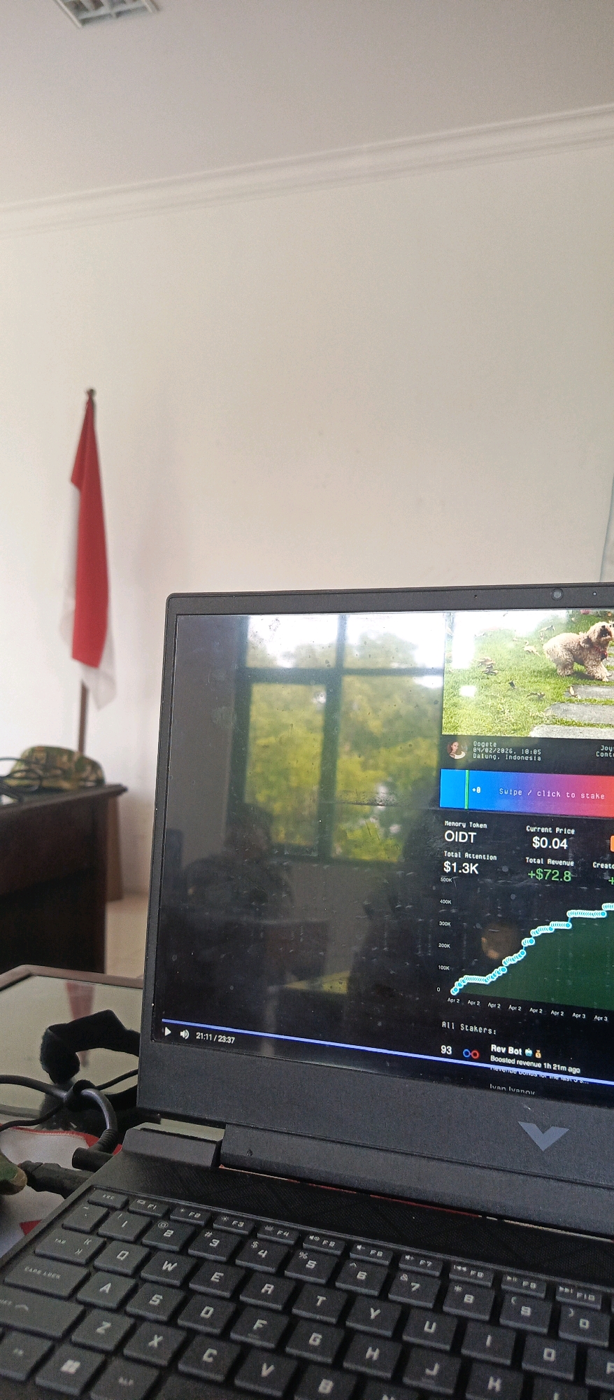 The image shows a laptop displaying a cryptocurrency trading interface. The screen shows a graph of "OIDE" token's price performance, with a current price of $0.04 and total revenue of +$72.8. The graph indicates a rising trend. There is also information about "Total Attention" being $1.3K. The interface includes a timestamp of "21:11 / 23:37", suggesting a video or playback is in progress. The background of the laptop screen shows a reflection of a window with greenery outside, and a small image of a dog on a grassy area.
In the background, a red and white Indonesian flag is visible on a pole. A desk with a military-style cap is also present to the left of the laptop. The room appears to be a well-lit indoor space with white walls and a ceiling. The overall scene suggests a work or study environment, possibly in a home office or a similar setting. The reference to "Datung, Indonesia" within the displayed text on the laptop screen indicates a potential location, but it is not definitively confirmed to be the city of Batu, Indonesia without further contextual information.