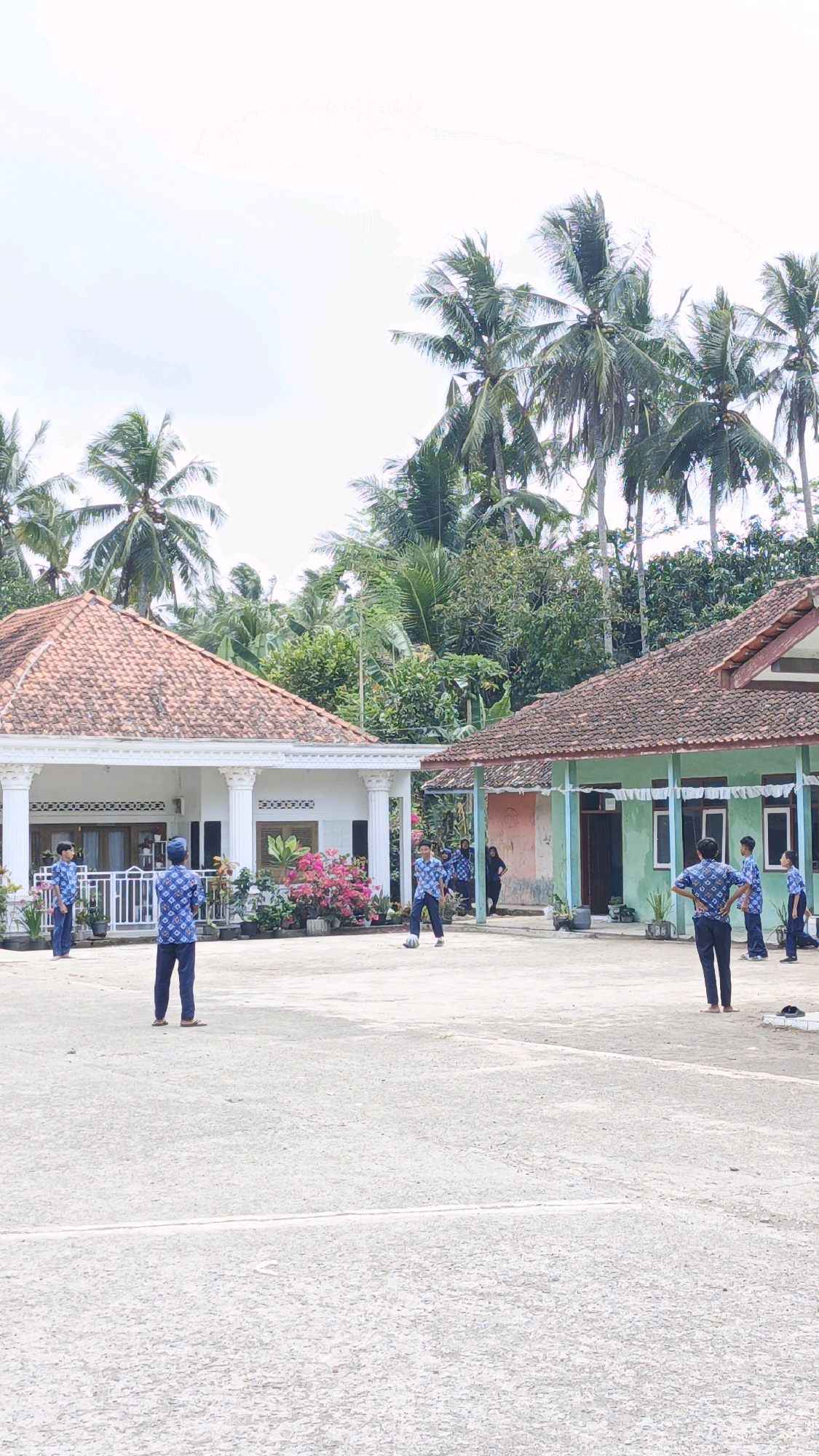 In Cibenda, Indonesia, a group of young males, some wearing matching blue patterned shirts and dark pants, are on an outdoor concrete court. Two individuals in the foreground are standing and facing each other, with one appearing to kick a white soccer ball. In the background, a building with a terracotta tiled roof and white columns is visible, adorned with potted plants. Adjacent to this, another building with a similarly tiled roof and a light green facade stands. Palm trees and dense green foliage surround the buildings and court, suggesting a tropical or subtropical environment. The sky is overcast with a pale, bright sky.