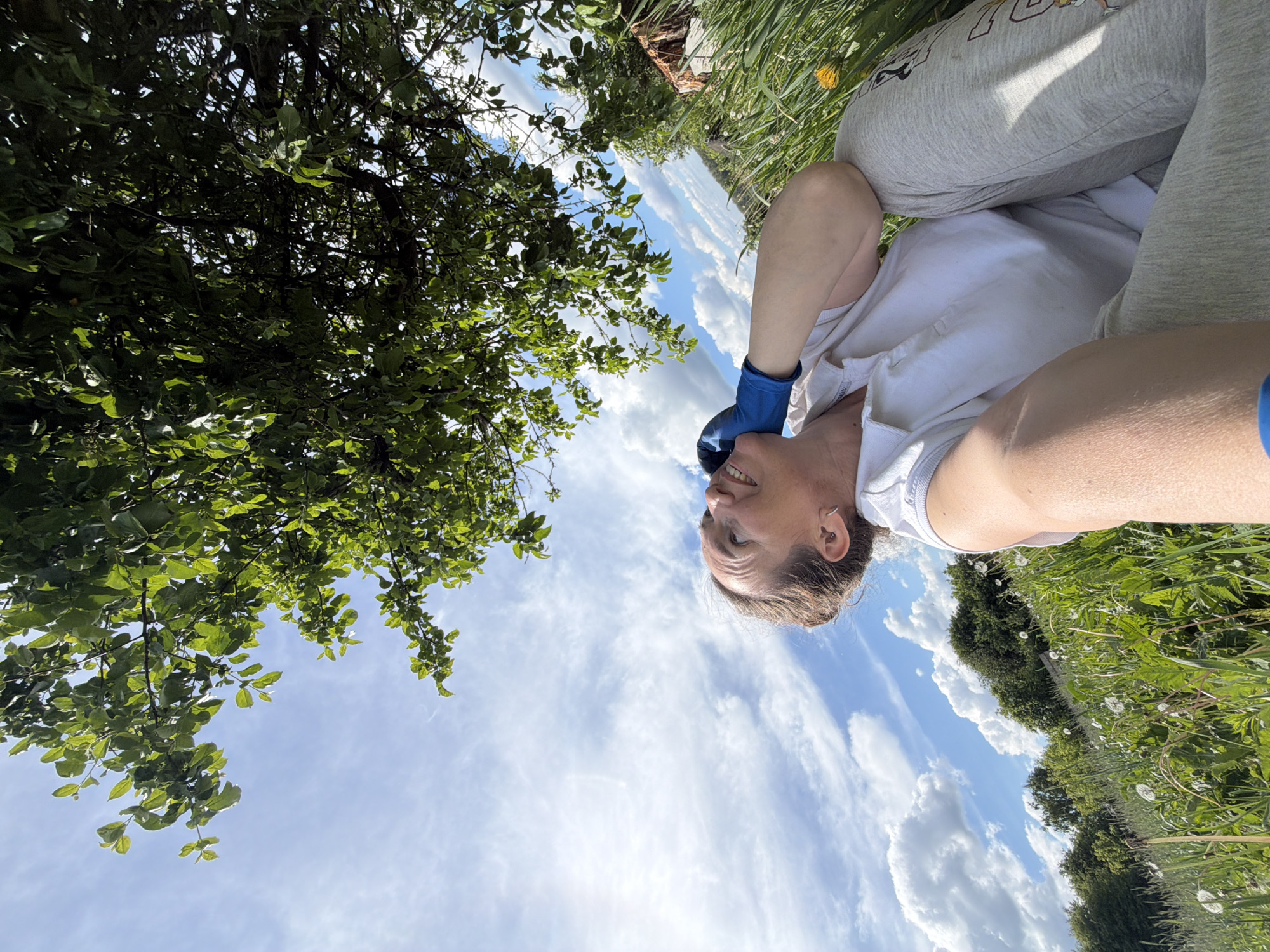 A low-angle outdoor photograph shows a person lying on their back in a grassy area, looking upwards towards the sky and tree canopy. The person, an adult, wears a white short-sleeved top and light gray sweatpants featuring a partially visible dark logo or text on the left thigh. Their right arm is bent with a blue glove on the hand, positioned near their chin. A subtle smile suggests a pleasant or relaxed mood.
The scene is dominated by bright green tree leaves and branches overhead on the left, with patches of bright blue sky and fluffy white clouds visible through the foliage and across the upper right. The ground is covered in tall green grass, interspersed with yellow dandelions and numerous white, wispy seed heads, indicative of late spring or early summer. In the distant background, a line of trees or bushes can be seen under the clear sky. The overall impression is one of peaceful outdoor relaxation or observation during bright daylight, possibly in a natural setting like a field or park in Olkhovka, Russia.