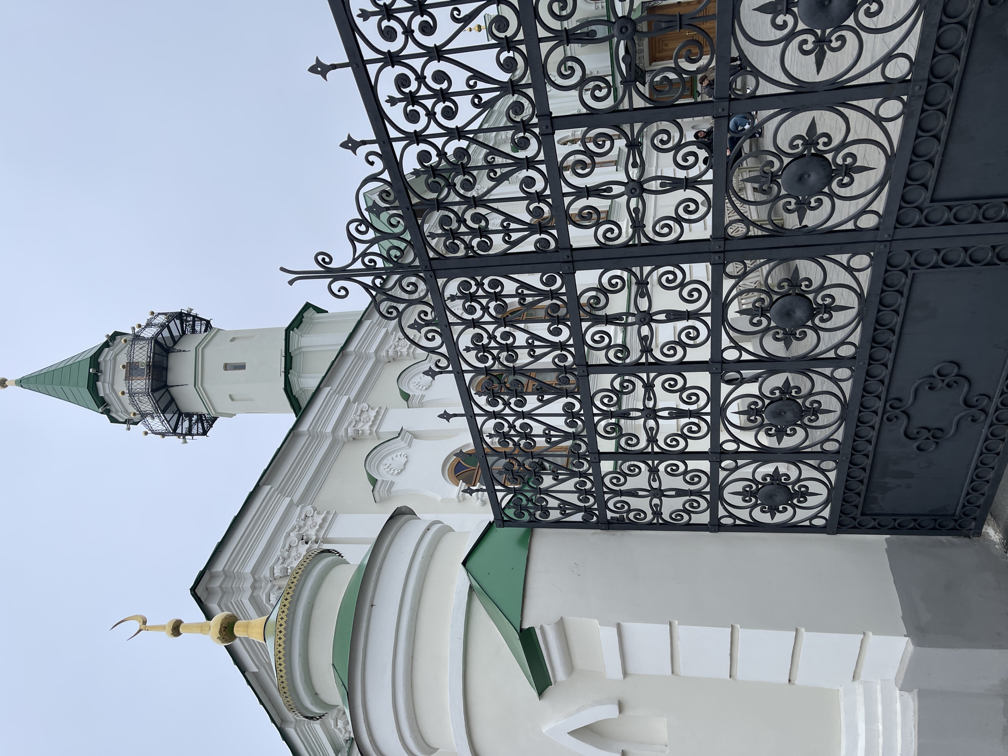 The image depicts a low-angle view of the Kul Sharif Mosque, located in Kazan, Russia, partially obscured by an ornate black metal fence. The mosque features white walls with green roofs and gold crescents atop its domes. A tall, slender minaret with a green roof is visible on the left side of the frame. The sky is overcast, suggesting a cloudy day. The fence occupies a significant portion of the foreground and right side of the image, and people can be seen in the background of the fence.