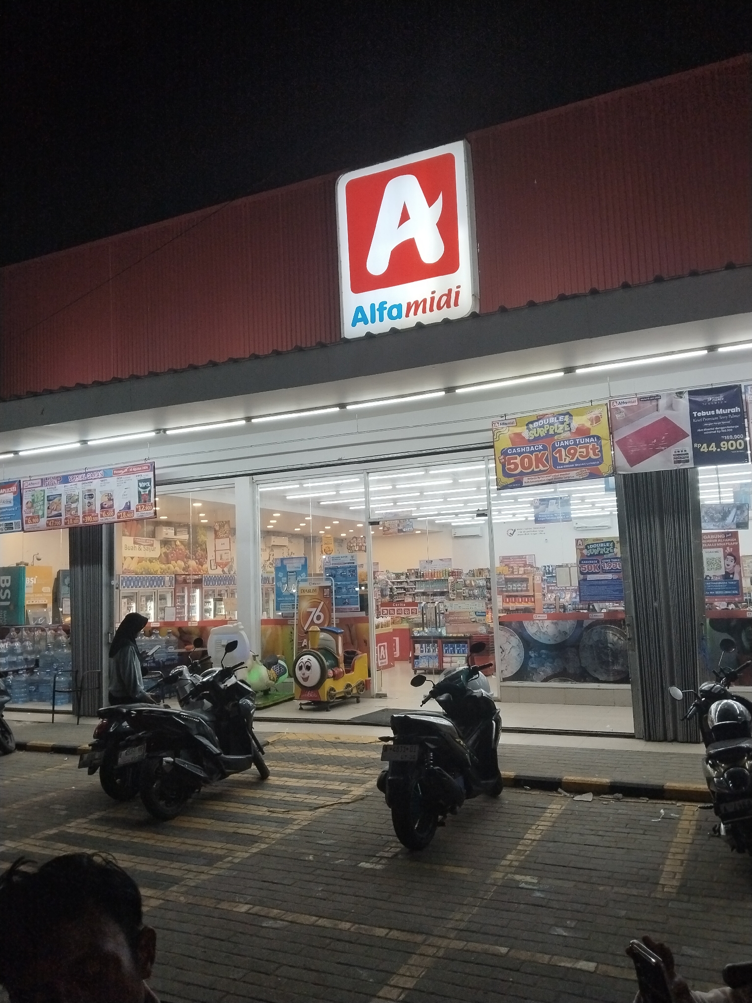 The image depicts the exterior of an Alfamidi convenience store in Carita, Indonesia, at night. A prominent illuminated sign with a red "A" and the text "Alfamidi" is visible at the top of the storefront. The storefront features large glass windows, revealing rows of brightly lit shelves stocked with products inside. Several promotional banners are displayed on the exterior glass.
In front of the store, a paved area with yellow lines marking parking spaces is visible. Three motorcycles are parked on this paved area. One person, wearing a dark hijab and a light-colored jacket, is standing near a motorcycle in front of the store entrance. The overall environment suggests a commercial setting in a town or city. The lighting indicates it is nighttime.