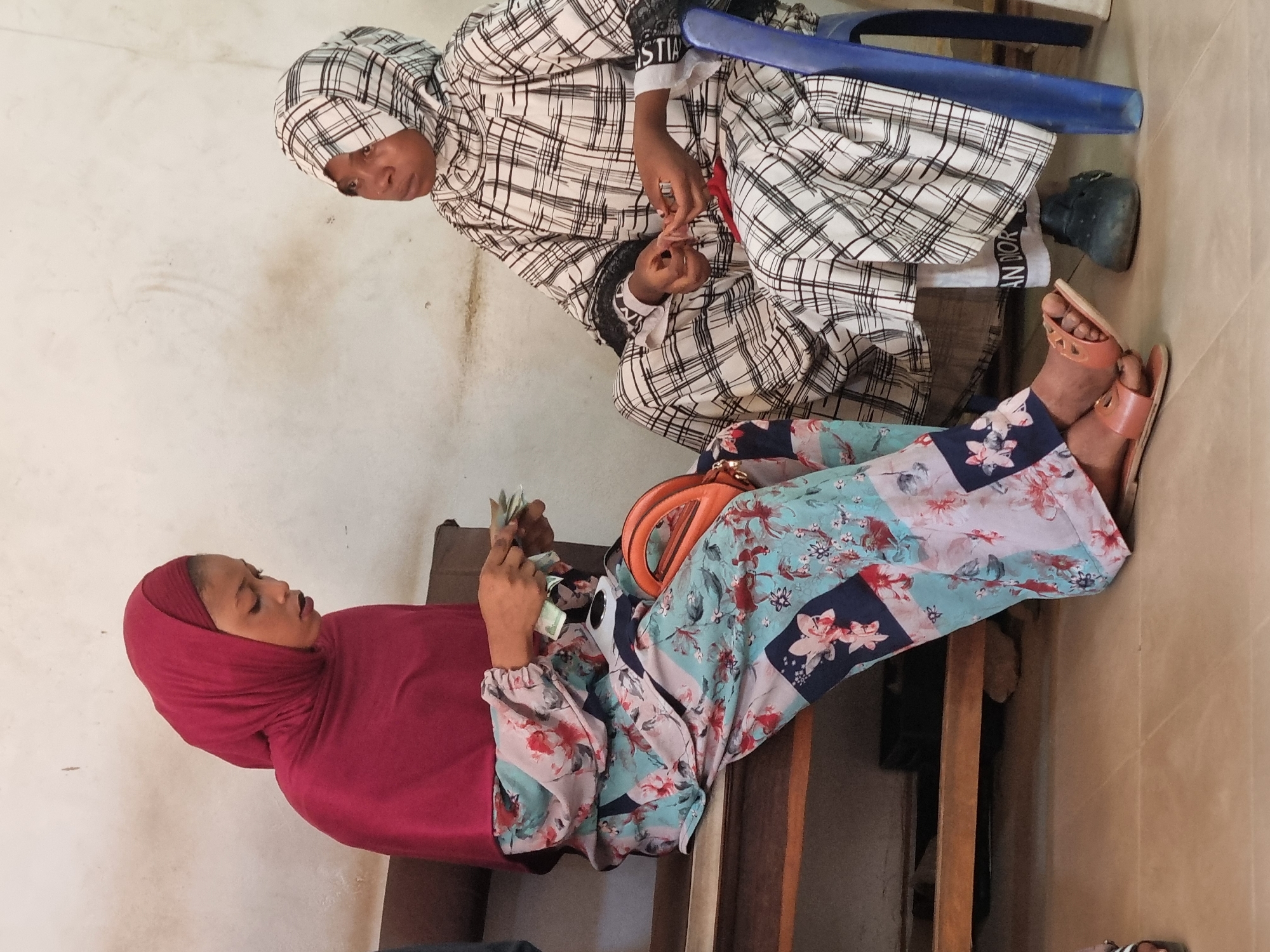 Two young women are sitting in what appears to be an indoor setting, possibly in Jos, Nigeria. One woman wears a burgundy hijab, floral patterned clothing, and sits on a wooden bench, counting money. An orange handbag rests next to her. The other woman is seated on a blue chair, wearing a checkered outfit and head covering, and has on a black boot with "CHRISTIAN DIOR" writing. The wall in the background is light-colored and appears somewhat worn.
