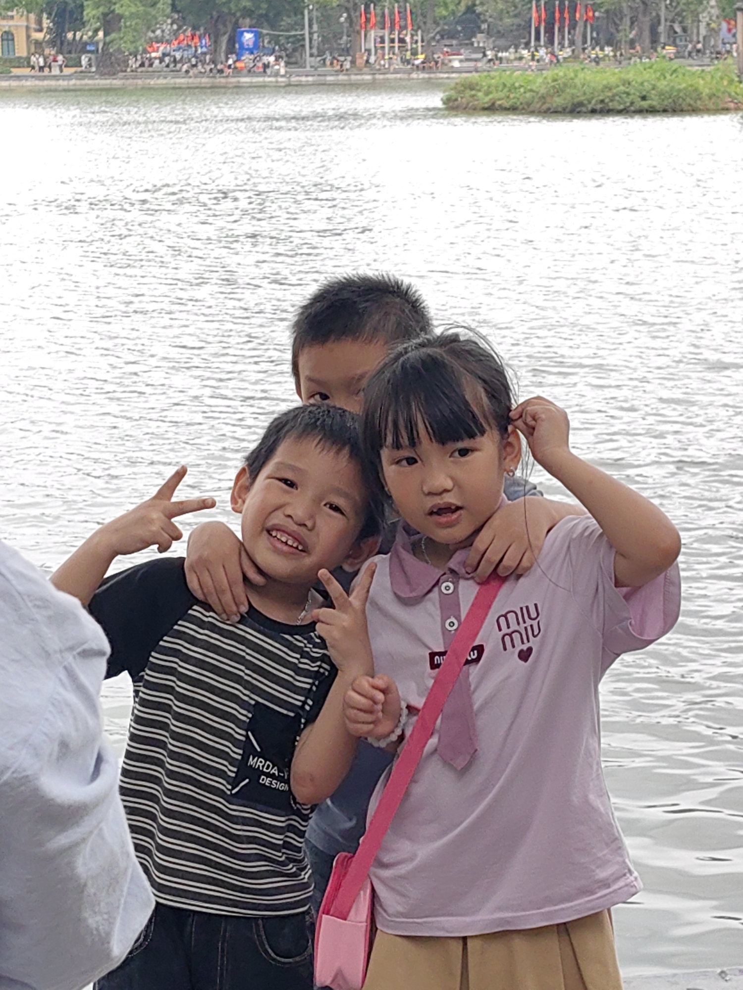 A medium shot captures three young children posing playfully near a body of water, likely a lake or river, in Hà Nội, Vietnam. The setting appears to be a bright, possibly overcast, daytime.
In the foreground, slightly to the left, a portion of an adult's light-colored clothing is visible. Centrally, a boy with a wide smile and dark hair, wearing a black and white striped t-shirt, makes peace signs with both hands. His left arm is around the shoulder of the girl to his right, and his right arm is around the shoulder of another boy partially obscured behind them. The striped t-shirt features text that reads "MRDA-X DESIGN" on a dark patch on the left sleeve/shoulder area.
To the right, a girl with dark hair and bangs, wearing a light pink collared polo shirt, adjusts something near her ear with her left hand. Her right hand is resting near her waist. Her shirt has a pink and white collar, two white buttons, and the brand name "MIU MIU" printed multiple times in purple, along with a small purple heart logo. She wears a pink cross-body strap, possibly for a small bag, which is partially visible at the bottom right. Her lower body is clad in a light brown pleated skirt. The third child, a boy, is mostly visible from the top of his head to his eyes, positioned behind the smiling boy and the girl, with his arm around the girl's shoulder.
The children are standing beside a calm body of water that fills the midground and most of the background. On the far bank, a small green, vegetated island is visible. Beyond it, there are trees, distant urban structures, and numerous small red flags arranged in a line, suggesting a festive or public area. People are scattered along the distant shoreline. The overall mood is cheerful and candid.