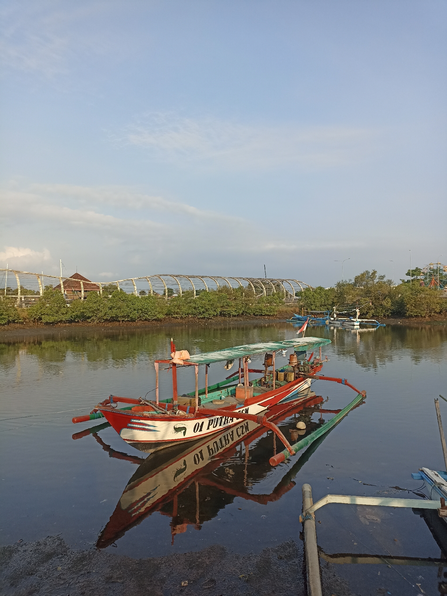 This is a photograph taken in Banyuwangi, Indonesia, likely near the Mangrove Planting and Edu Park. The image captures a serene waterscape with several boats docked or moored. In the foreground, a traditional Indonesian fishing boat with a red and white hull and a green canopy is prominently featured. The boat's reflection is clearly visible on the calm water. In the background, a distinctive white, arching structure, characteristic of the park's entrance or a similar architectural feature, spans across the scene. Lush green mangrove trees line the opposite bank of the waterway. The sky is a pale blue with scattered clouds, suggesting it is either early morning or late afternoon. The overall impression is one of peaceful, natural beauty with a touch of local culture evident in the boats and the surrounding environment.