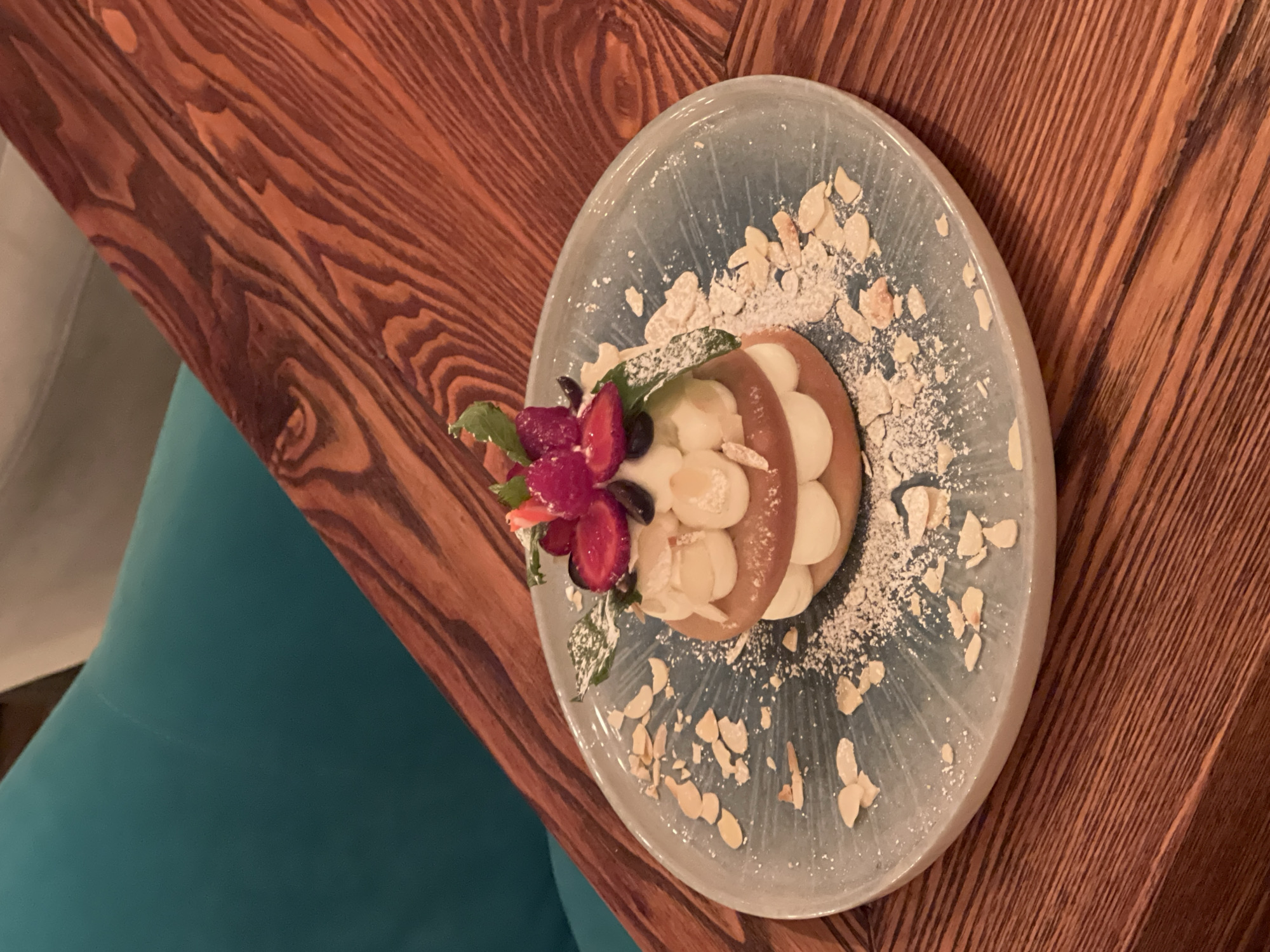 A delicately prepared dessert sits on a light blue ceramic plate, which is placed on a rich, dark wooden table. The dessert appears to be a layered cake or pastry, adorned with fresh berries, possibly raspberries and strawberries, blueberries, and fresh mint leaves. Sliced almonds and a dusting of powdered sugar are scattered on the plate around the dessert. The background shows a portion of a teal upholstered chair and the same wooden table. The scene is likely indoors, potentially in a cafe or restaurant. The context provided states the location is Novosibirsk, Russia.