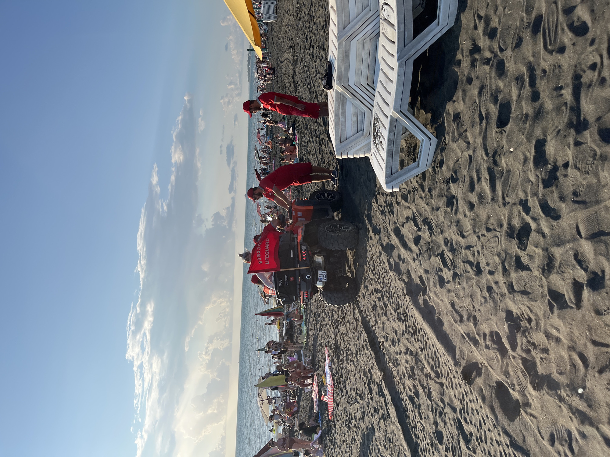 The image depicts a crowded beach in Ureki, Georgia, on a sunny day with a blue sky and scattered clouds. In the foreground, dark sand is visible with tire tracks and footprints. Several white lounge chairs are stacked in the middle ground. A red ATV, equipped with a red flag that reads "LIFEGUARD" in English and Georgian, is parked on the sand. Two lifeguards in red uniforms stand near the ATV. The background reveals the sea with gentle waves, and a multitude of beachgoers spread out on the sand, some under umbrellas and parasols. The overall scene conveys a lively and popular beach environment.