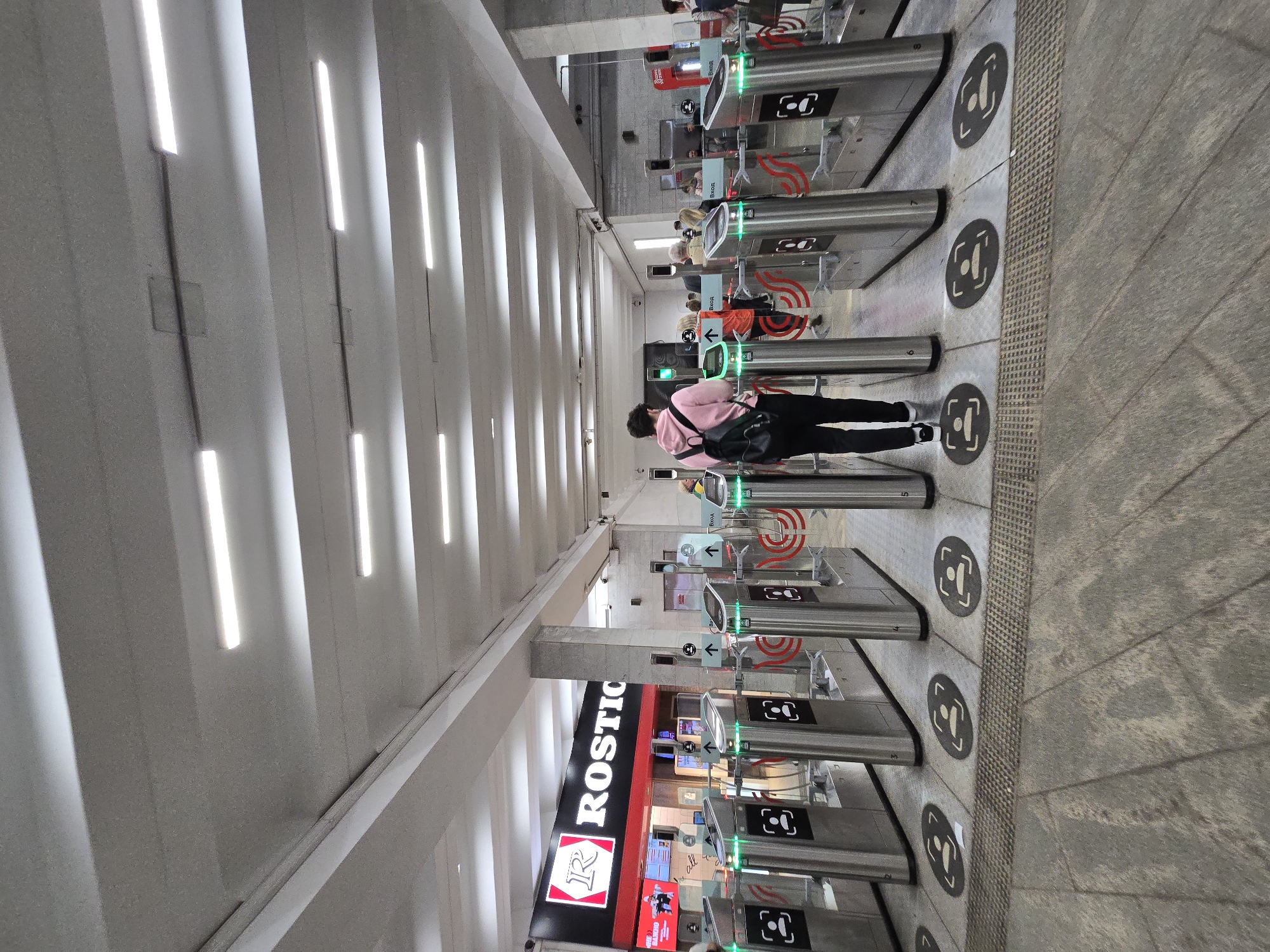 The image depicts the entrance of a metro station in Moscow, Russia. A person wearing a pink jacket and black pants is walking through a turnstile. Several turnstiles are aligned in rows with green lights indicating passage. The station interior features a ceiling with parallel rows of recessed linear lighting. On one side, a commercial sign for "ROSTIC'S" is visible. The floor is grey and patterned. Circular directional icons are painted on the floor next to the turnstiles.