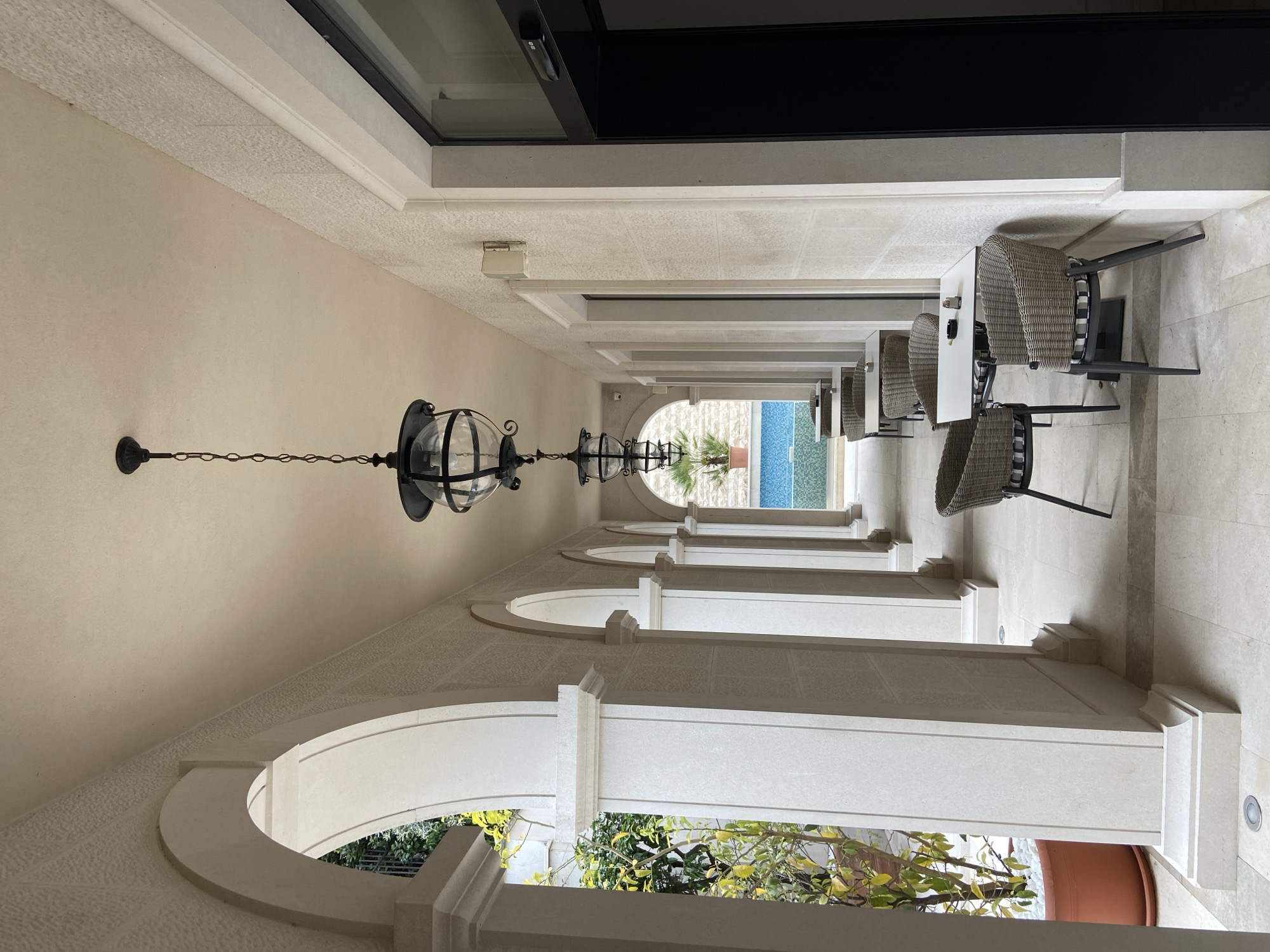 This image captures a long, elegant outdoor corridor or colonnade, characteristic of a high-end resort or hotel in Budva, Montenegro. The perspective looks down the length of the corridor, leading the eye towards a bright outdoor area.
On the left side of the corridor, a series of large, cream-colored arched openings frame views of lush green foliage and potted plants, suggesting an adjacent garden or courtyard. On the right, a more solid wall features recessed architectural panels and supports several outdoor dining arrangements. Each setting includes a small, white square table accompanied by two modern wicker-style chairs with dark cushions, inviting guests to sit and relax.
The ceiling of the corridor is adorned with multiple traditional, black wrought-iron hanging lanterns, each featuring a spherical glass enclosure around a light source. The floor and structural elements are constructed from light-colored, possibly beige or off-white, stone or tile, contributing to a sense of spaciousness and classic elegance.
At the far end of the corridor, through a final large archway, a vibrant blue swimming pool is visible. The pool features a distinctive mosaic tile pattern in varying shades of blue and green, and a potted palm tree stands beside it, hinting at a sunny and serene leisure area.
The overall architectural style is stately and Mediterranean, with clean lines, arches, and stone finishes. The bright, natural illumination suggests it is daytime, likely mid-morning or afternoon, given the lack of harsh shadows. There are no people present in the scene, and no visible text. The setting evokes a sense of peaceful luxury and an inviting atmosphere.